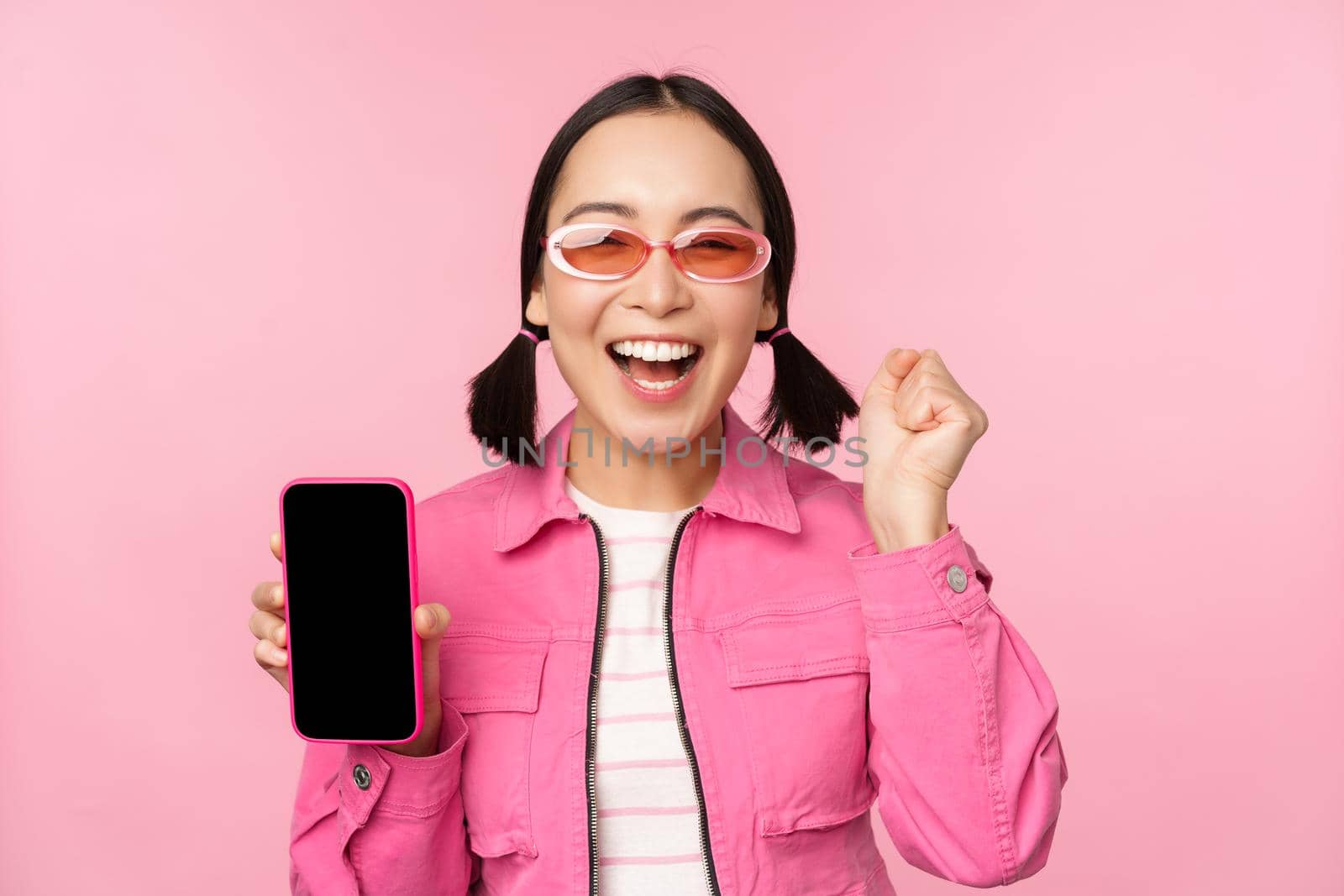 Excited asian girl laughs and smiles, shows mobile phone screen, smartphone application, standing over pink background by Benzoix