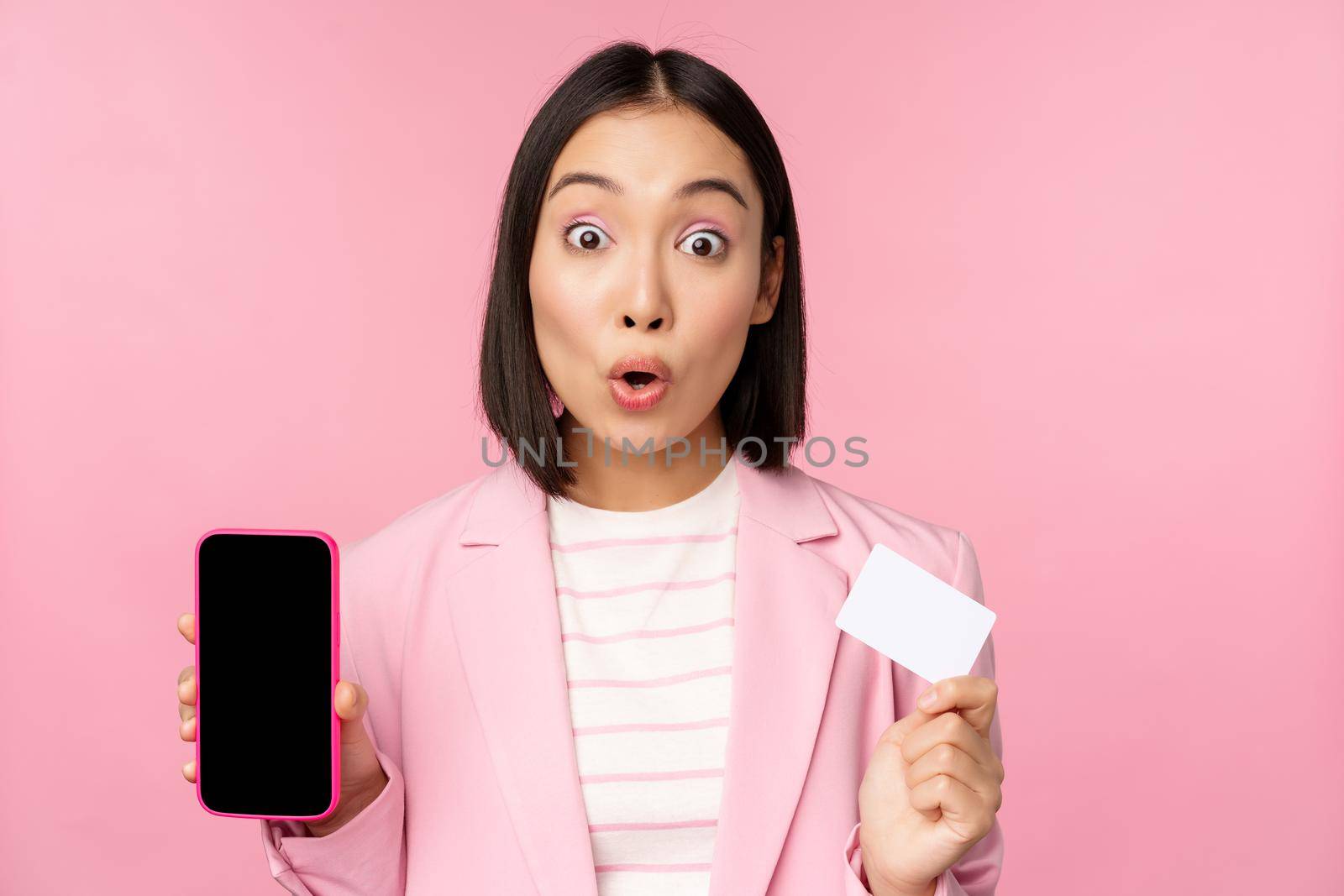 Enthusiastic asian businesswoman showing mobile phone screen and credit card, looking amazed at camera, standing over pink background.