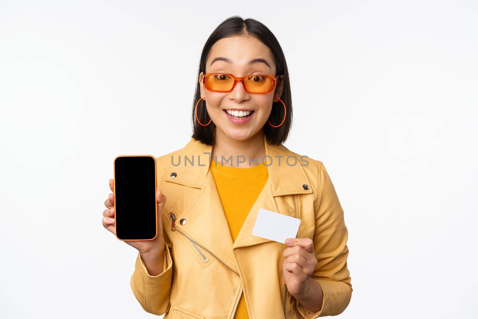 Online shopping and people concept. Stylish asian woman showing mobile phone screen and credit card, smartphone application, standing over white background by Benzoix