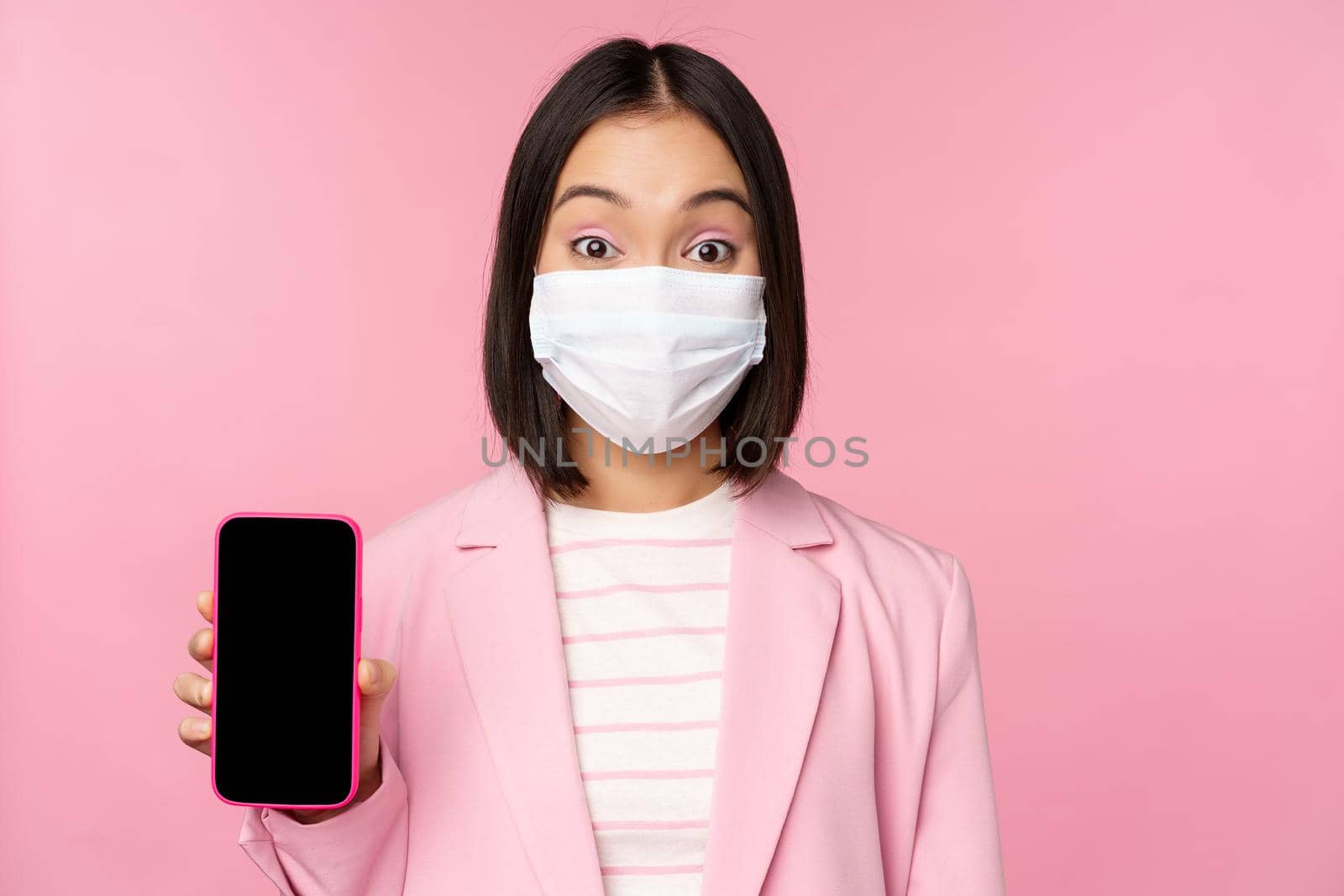 Portrait of smiling korean saleswoman in medical face mask, business suit, showing smartphone screen, standing over pink background.
