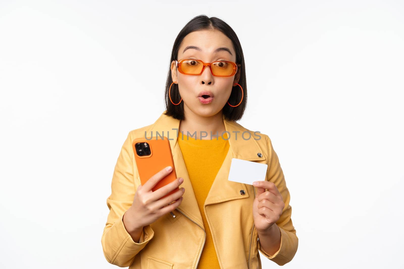 Online shopping. Stylish asian female model in sunglasses, holding credit card and mobile phone, smiling happy, standing over white background. Copy space