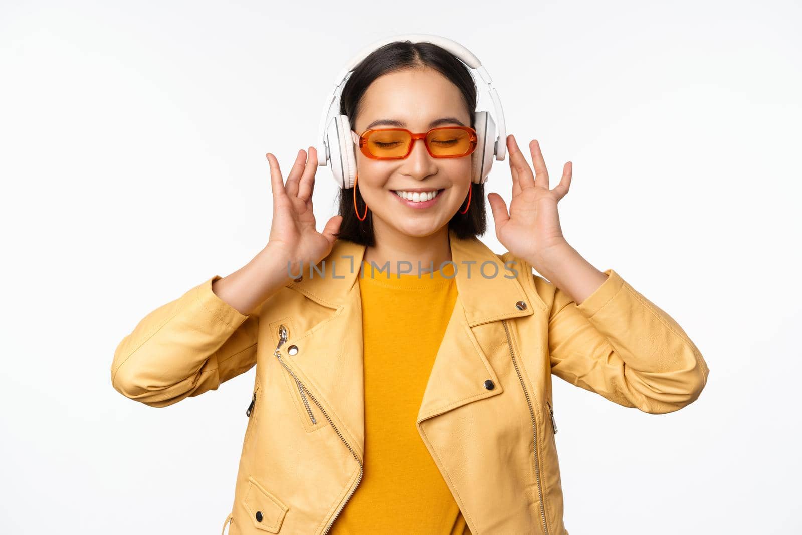 Beautiful asian girl, laughing happy, listening music in headphones, standing over white background. Copy space