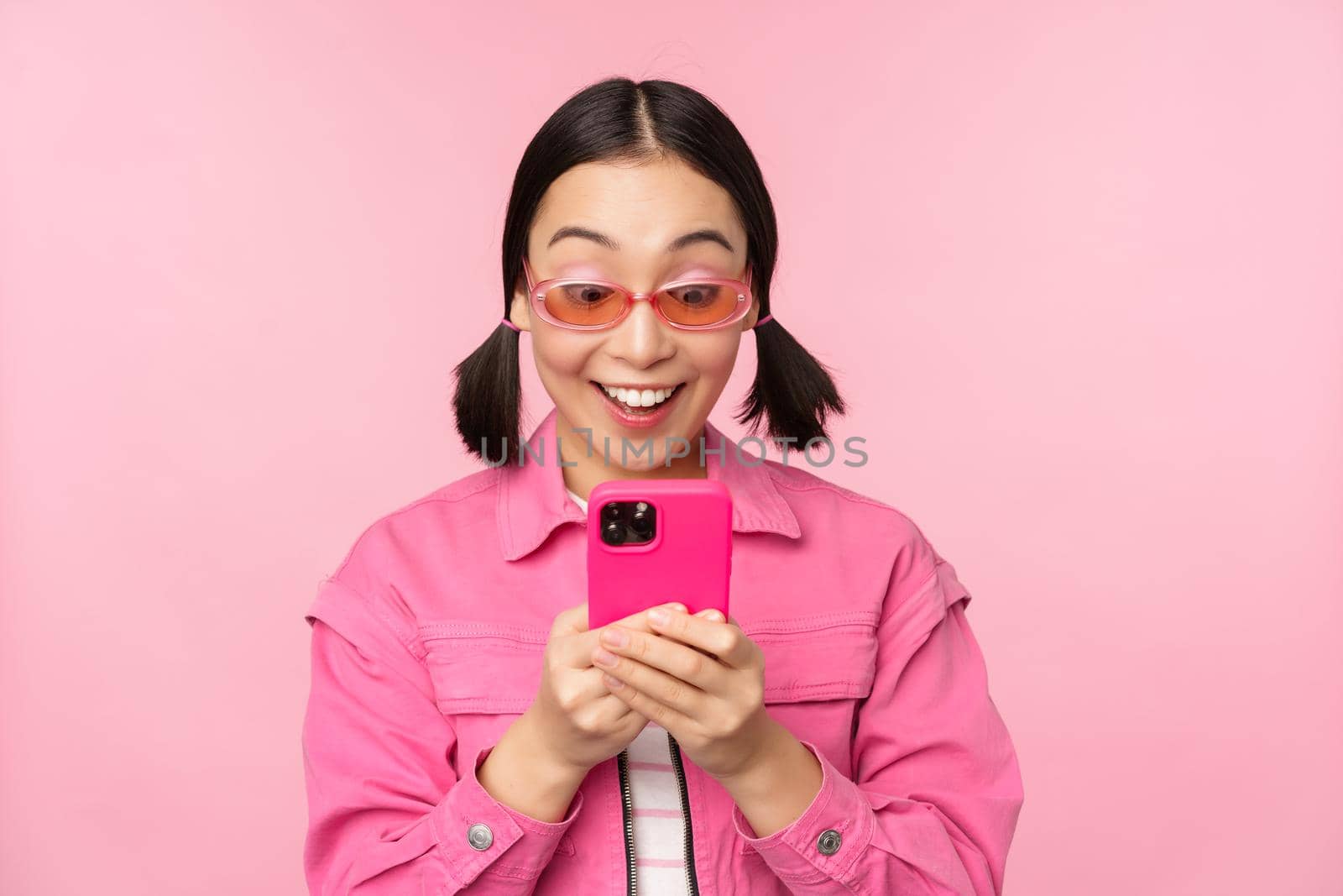 Portrait of asian girl in sunglasses using smartphone. Woman looking at mobile phone, browsing in app, standing over pink background by Benzoix