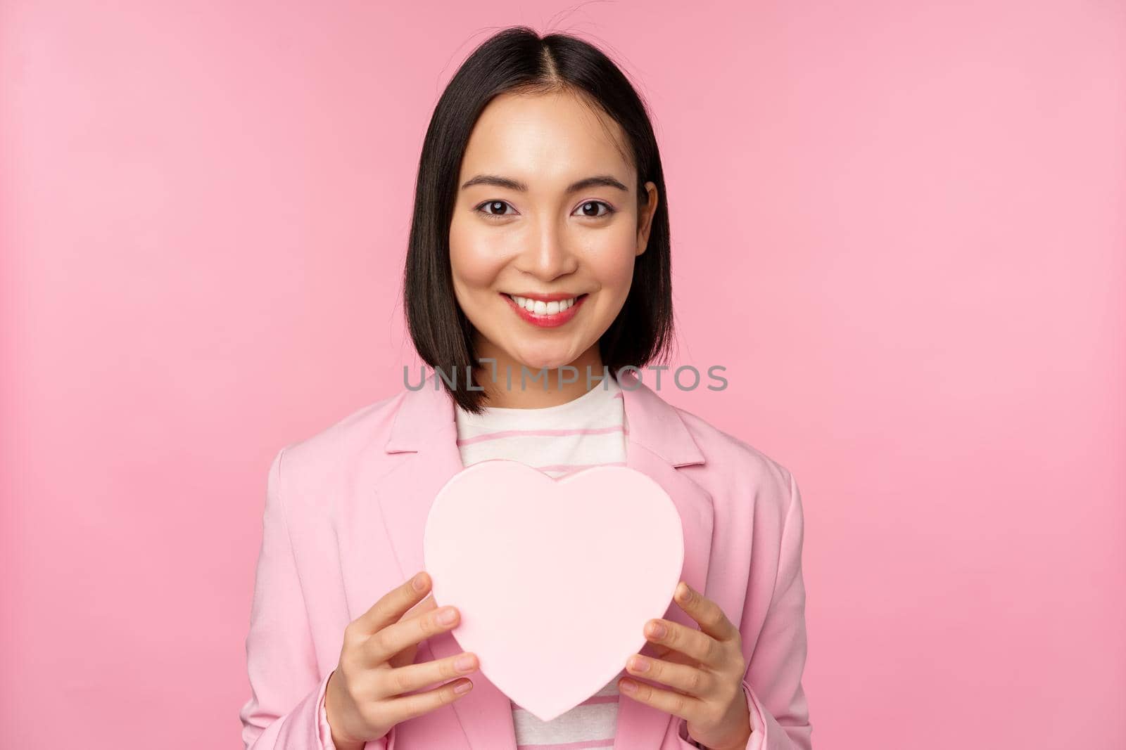 This is for you. Romantic cute asian corporate woman, girl in suit, showing heart-shaped box with gift, standing over pink background by Benzoix