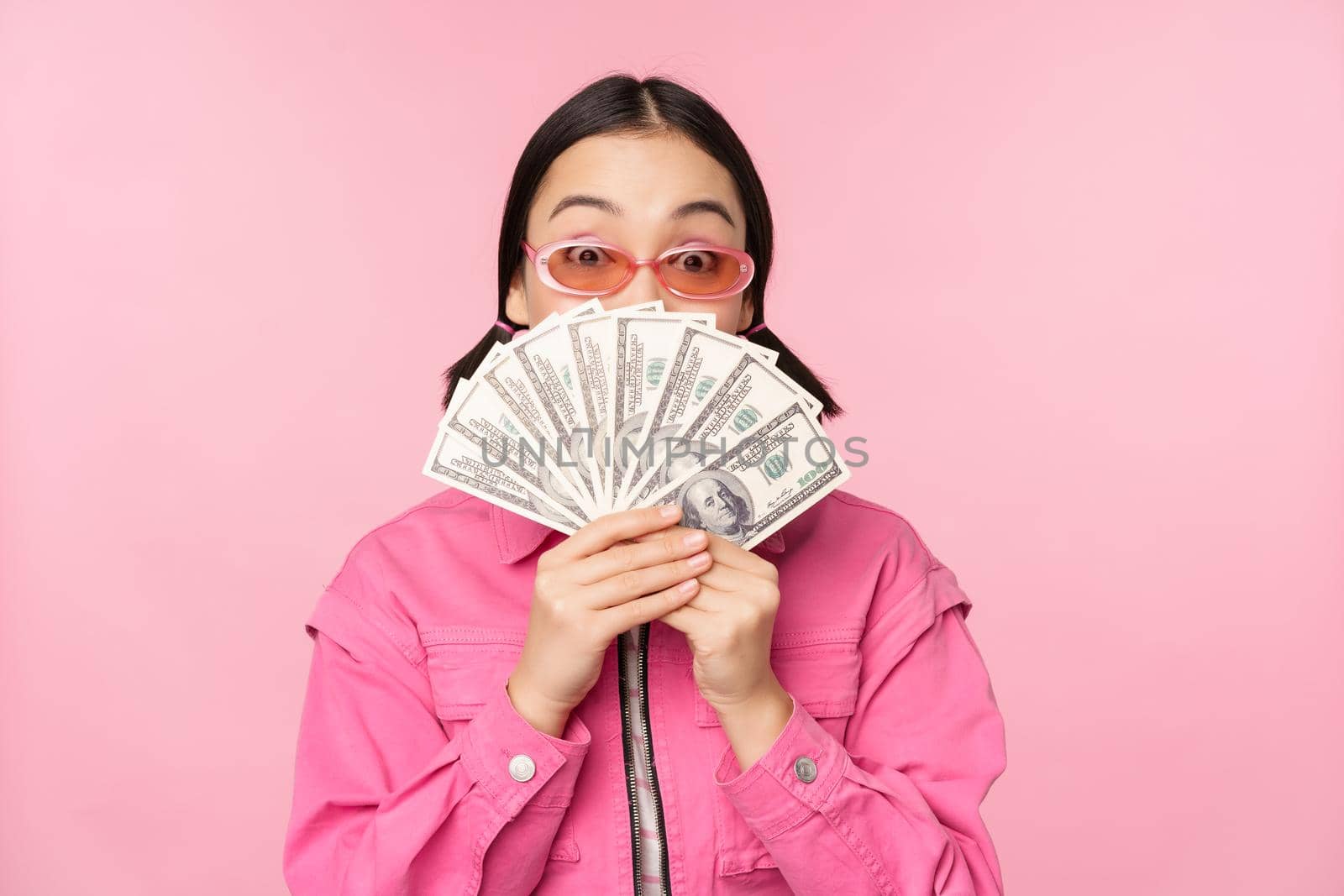 Beautiful korean woman in sunglasses, showing dollars, money cash, smiling pleased, concept of fast loans, microcredit and payment, standing over pink background.