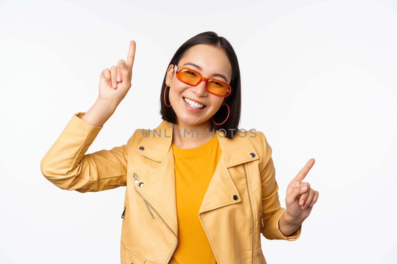 Smiling asian brunette woman in sunglasses, pointing fingers sideways, left and right, showing variants, laughing and dancing, wearing sunglasses, white background.