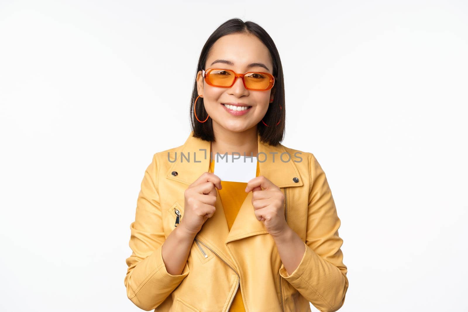 Portrait of smiling korean female model in sunglasses, showing credit card, standing over white background by Benzoix