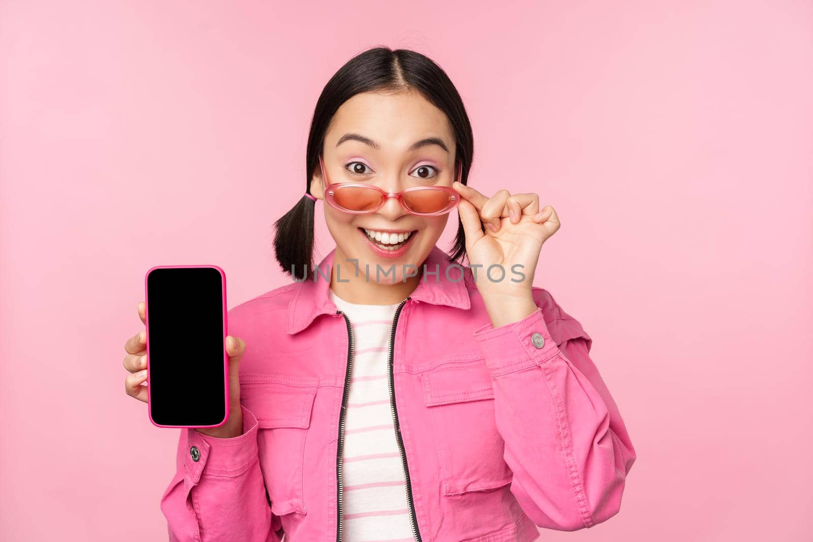 Portrait of asian girl showing mobile phone screen, reacting surprised, standing over pink background. Copy space