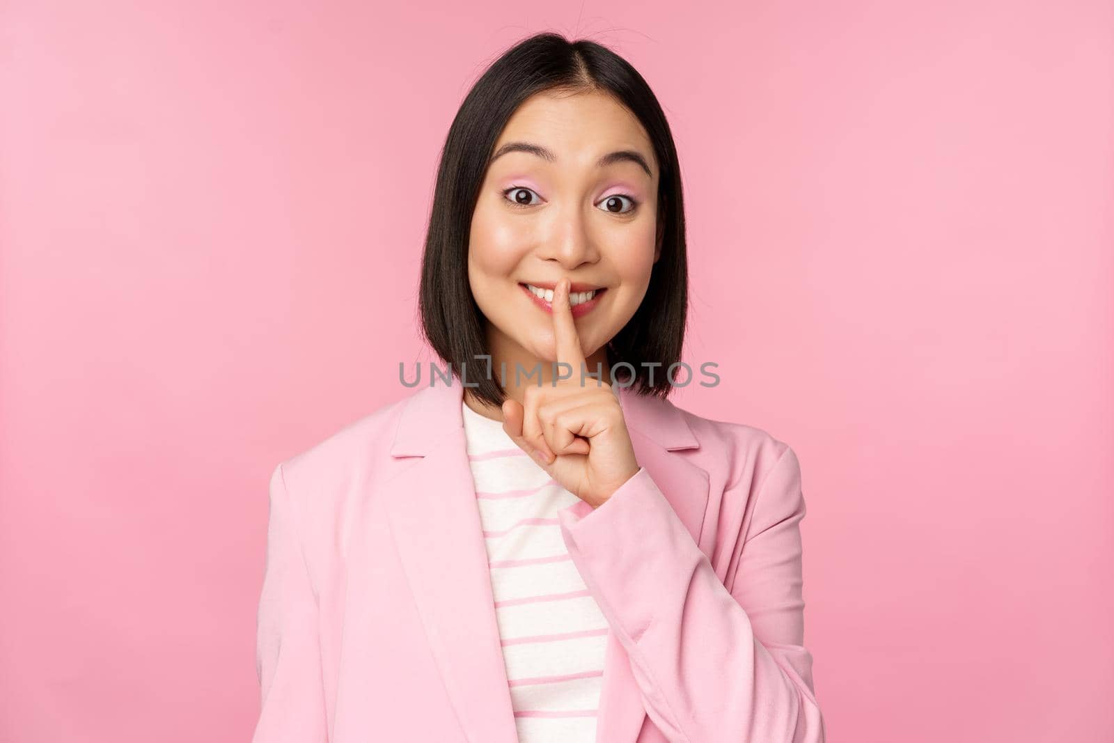 Hush, taboo concept. Portrait of asian businesswoman showing shush gesture, shhh sign, press finger to lips, standing over pink background in suit.