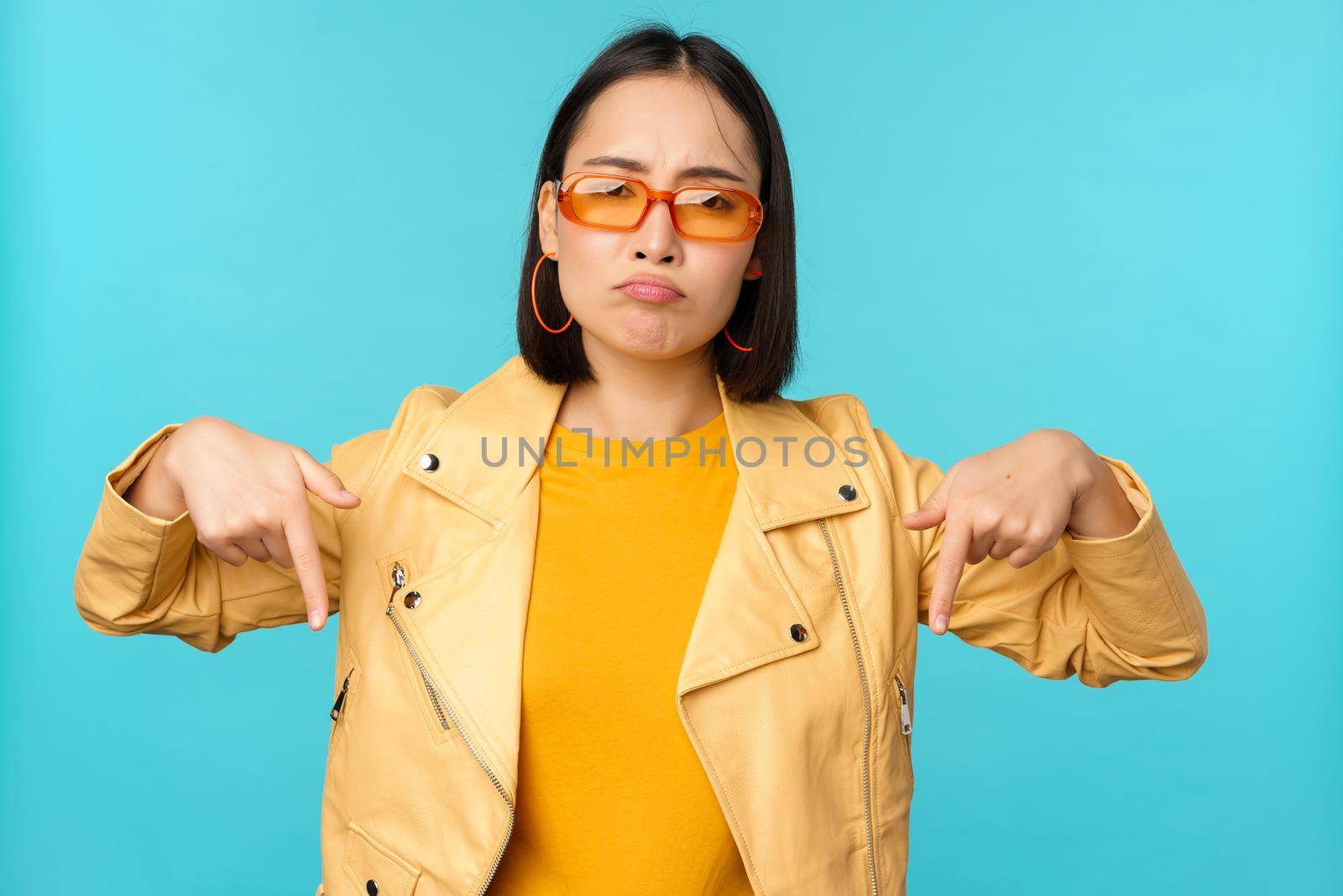 Sad and gloomy chinese girl in sunglasses, points fingers down and grimaces, looks miserable and disappointed, emotion of regret, standing over blue background.