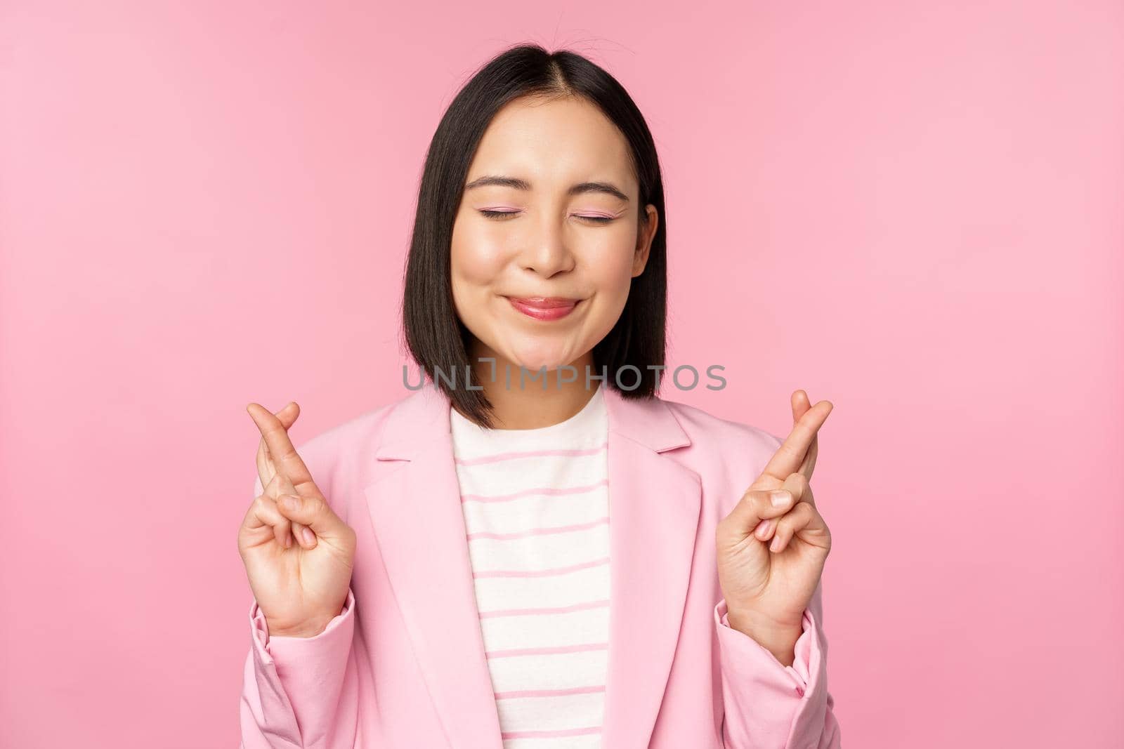 Happy lucky businesswoman, asian corporate lady wishing, making wish, hoping for smth and praying, standing in suit over pink background.