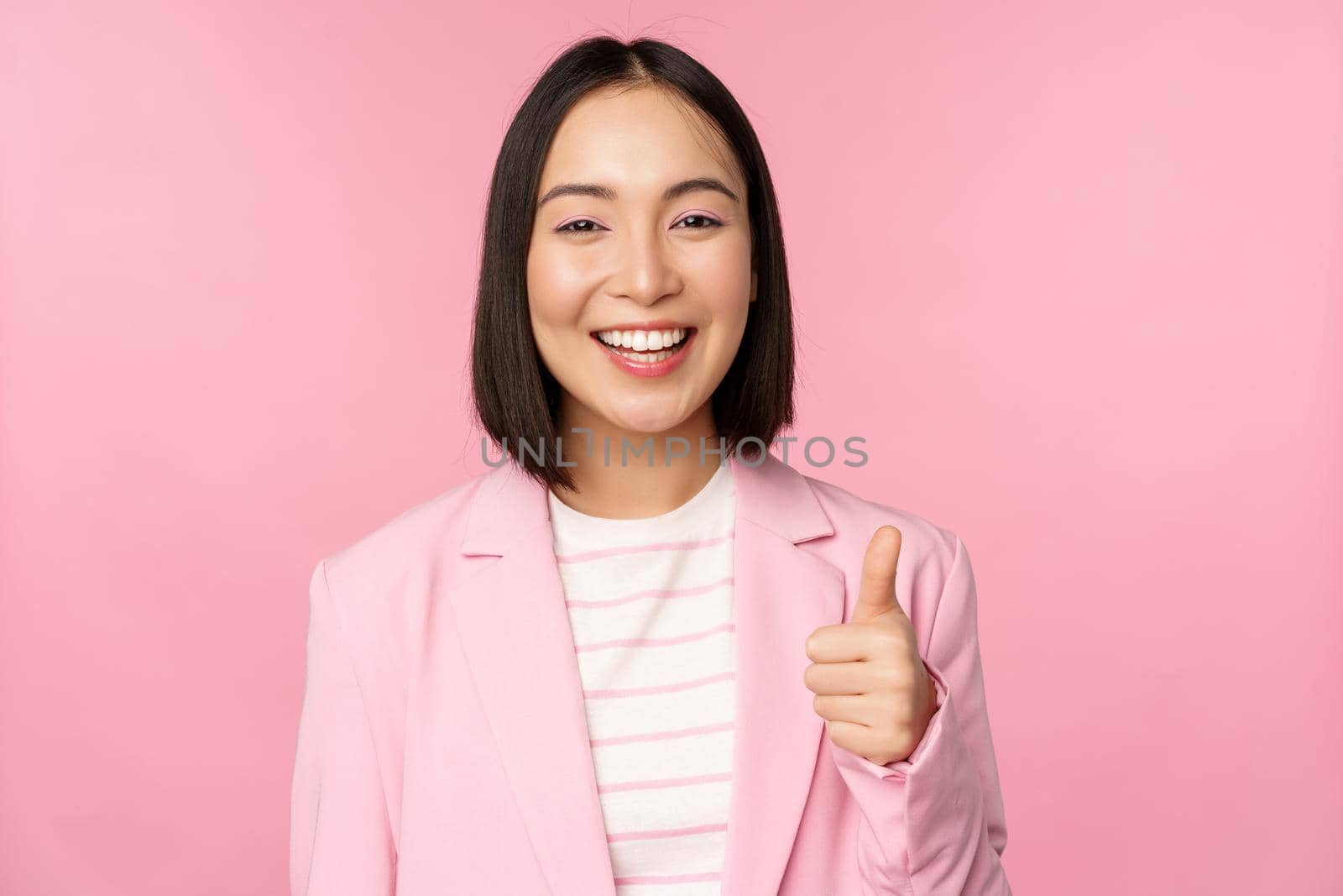 Portrait of asian businesswoman smiling satisfied, showing thumbs up, praise, like and approve, standing in suit over pink background by Benzoix