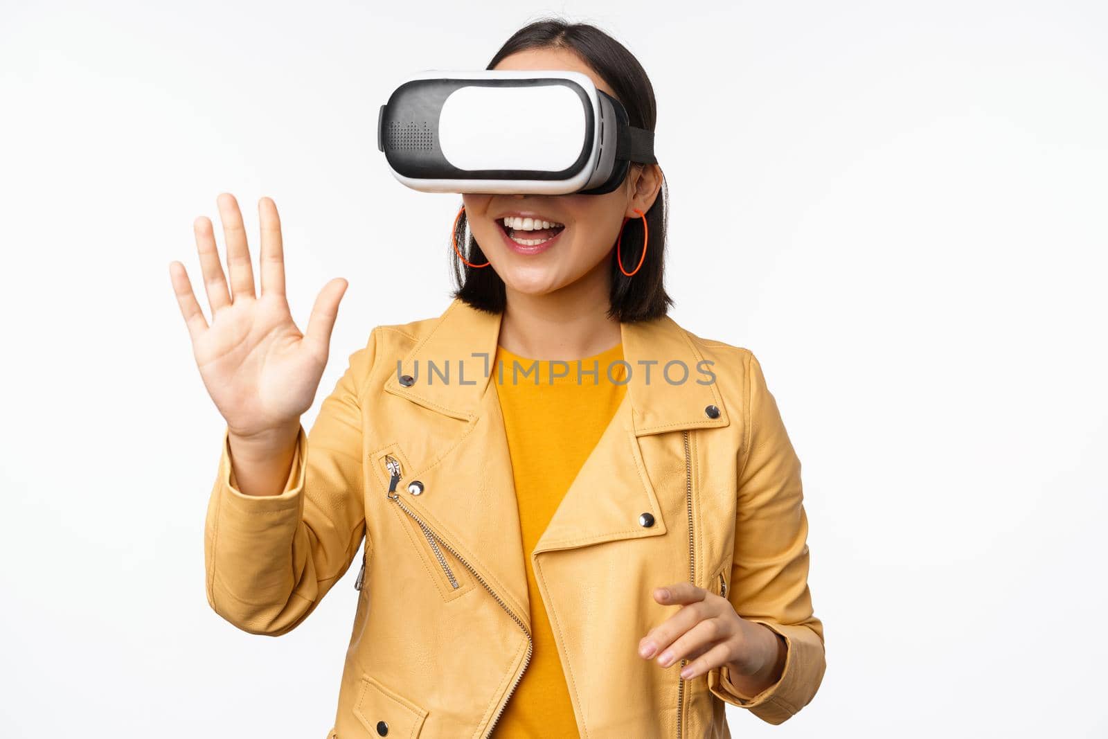 Happy asian girl in VR headset, saying hello in virtual reality chat, using special glasses, standing over white background, waving hand, saying hello.