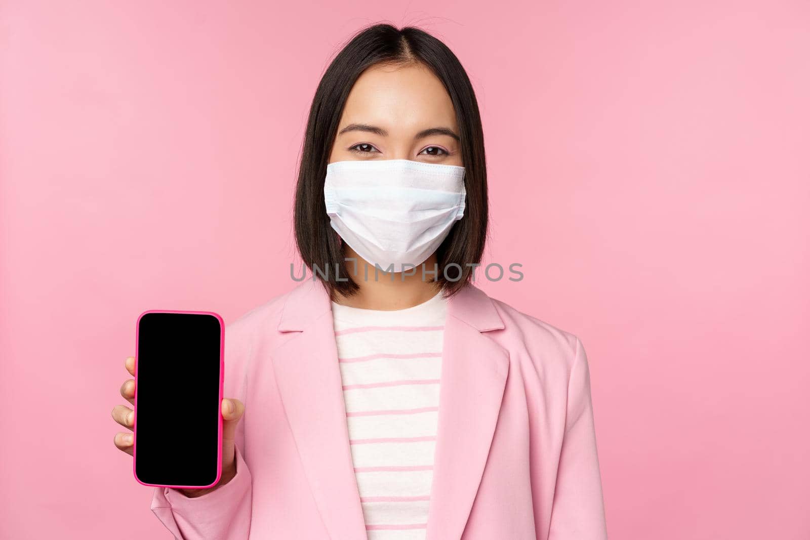 Portrait of smiling korean saleswoman in medical face mask, business suit, showing smartphone screen, standing over pink background by Benzoix