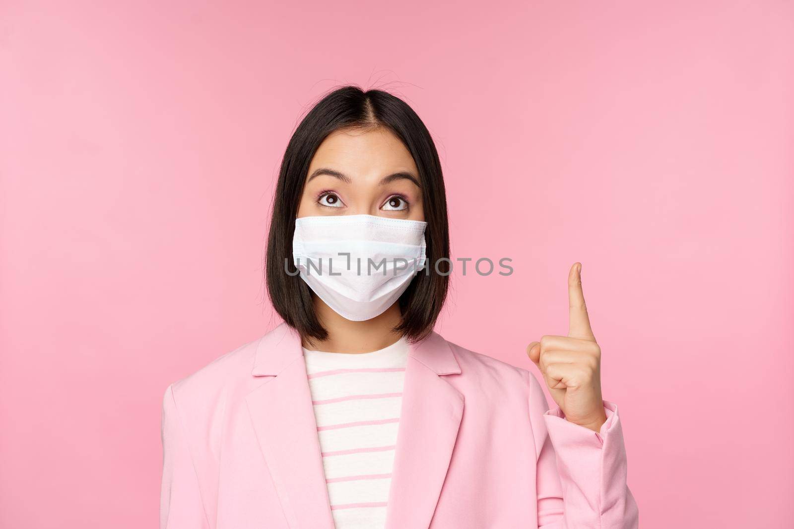 Close up portrait of businesswoman, asian lady in office suit and medical face mask, looking and pointing up, showing company logo or banner on top, pink background by Benzoix