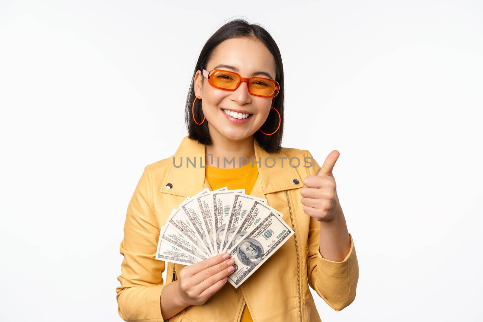 Stylish smiling asian girl holding money cash, showing dollars and celebrating, standing over white background. Copy space