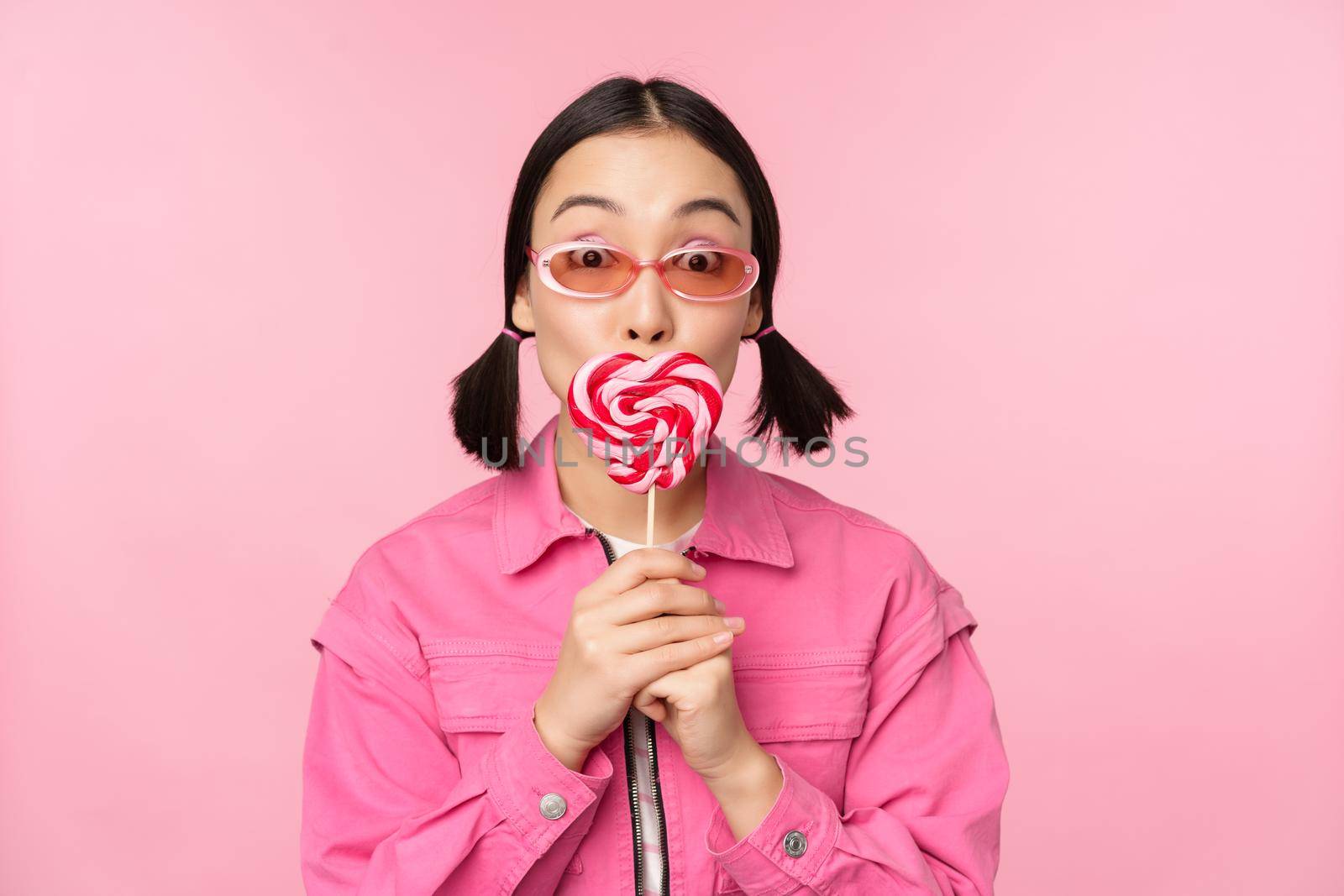 Stylish korean girl licking lolipop, eating candy and smiling, standing in sunglasses against pink background.