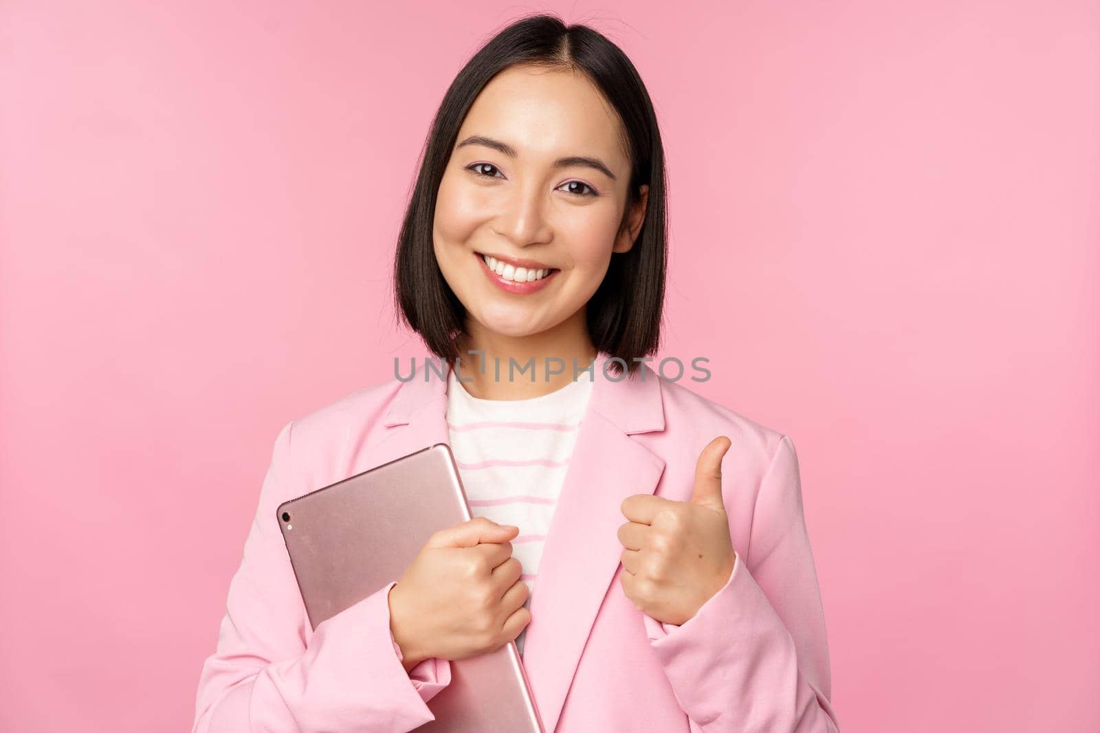 Portrait of corporate woman, girl in office in business suit, holding digital tablet, showing thumbs up, recommending company, standing over pink background.