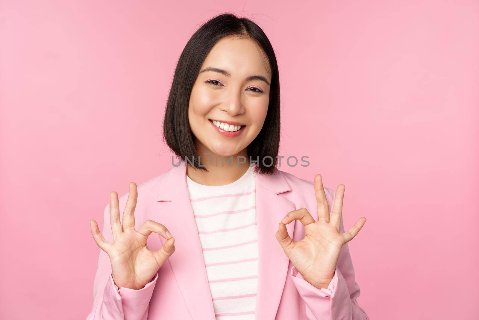 Okay, excellent. Businesswoman in corporate suit, showing ok, approval gesture, recommending smth, give positive feedback and smiling pleased, posing over pink background by Benzoix