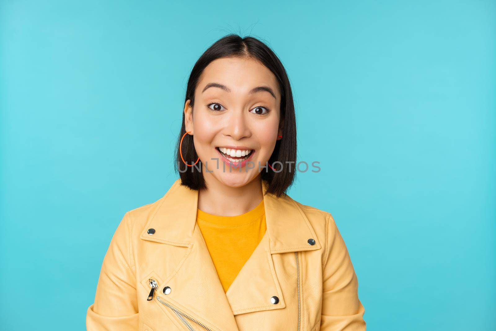 Close up portrait of asian girl looks surprised at amazed, wow face expression, stands over blue background.