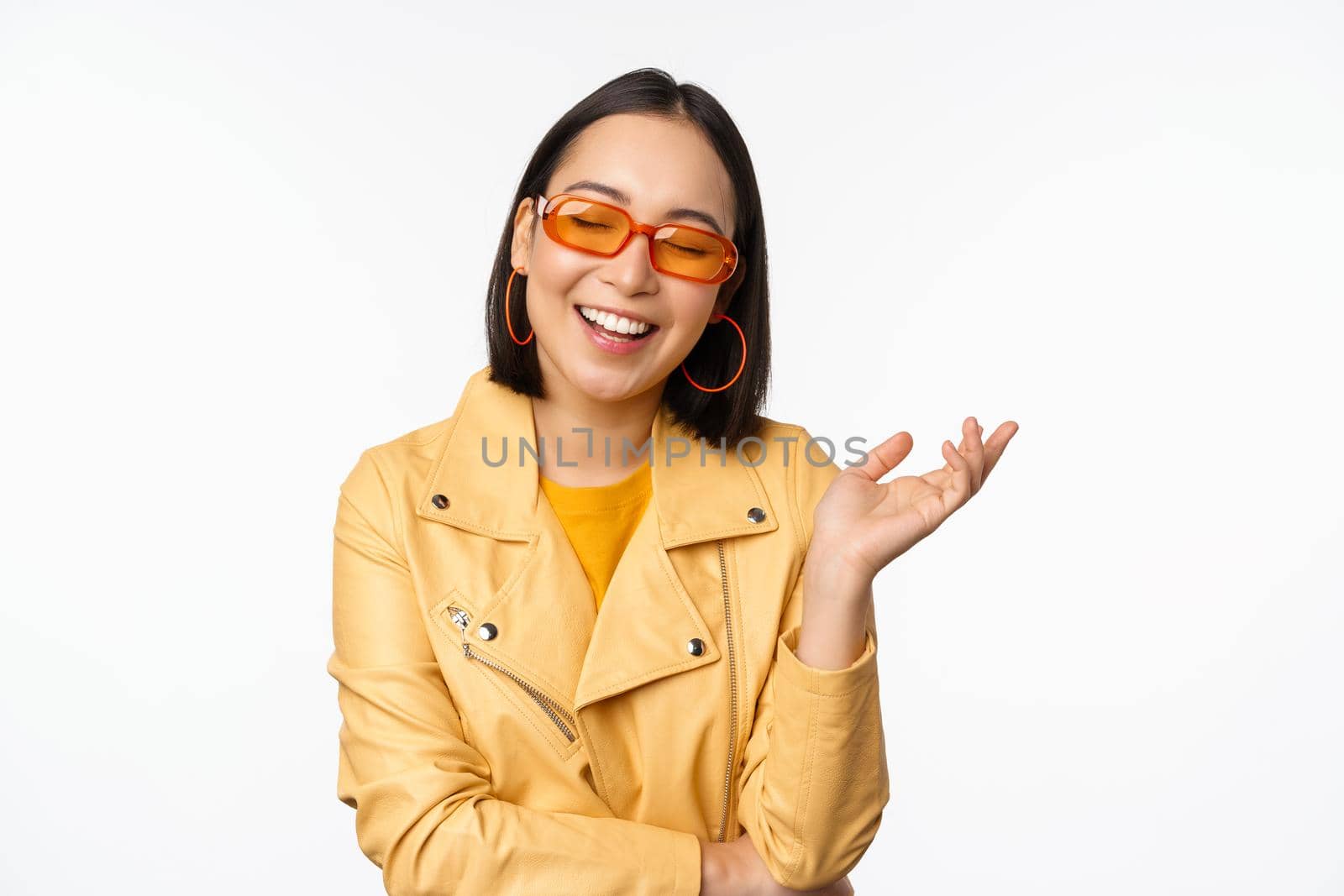 Stylish, modern korean girl in sunglasses and yellow coat, laughing and smiling, happy normal face expression, posing against white studio background.