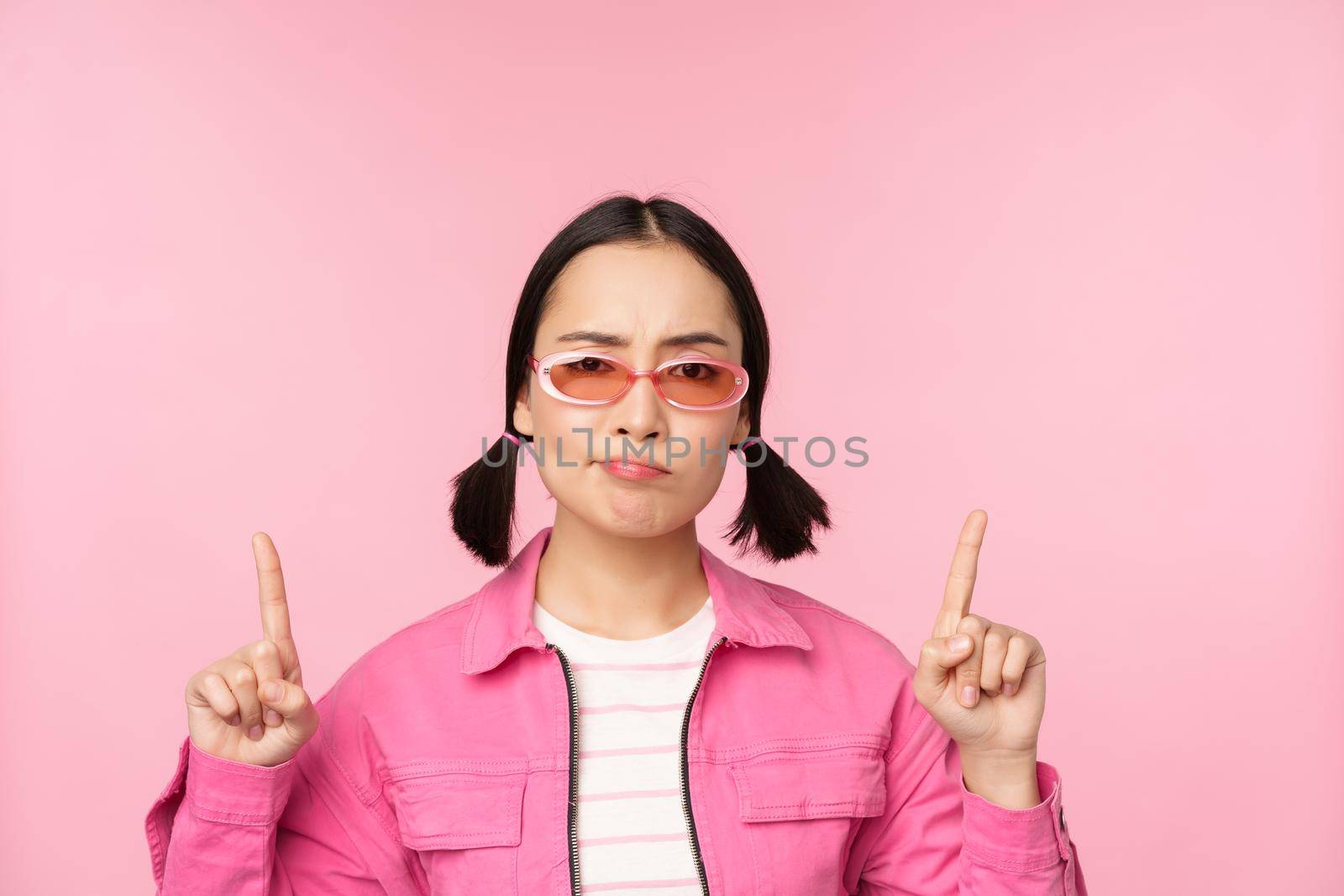 Close up of stylish korean girl, looking perplexed, pointing fingers up, showing advertisement, standing over pink background.