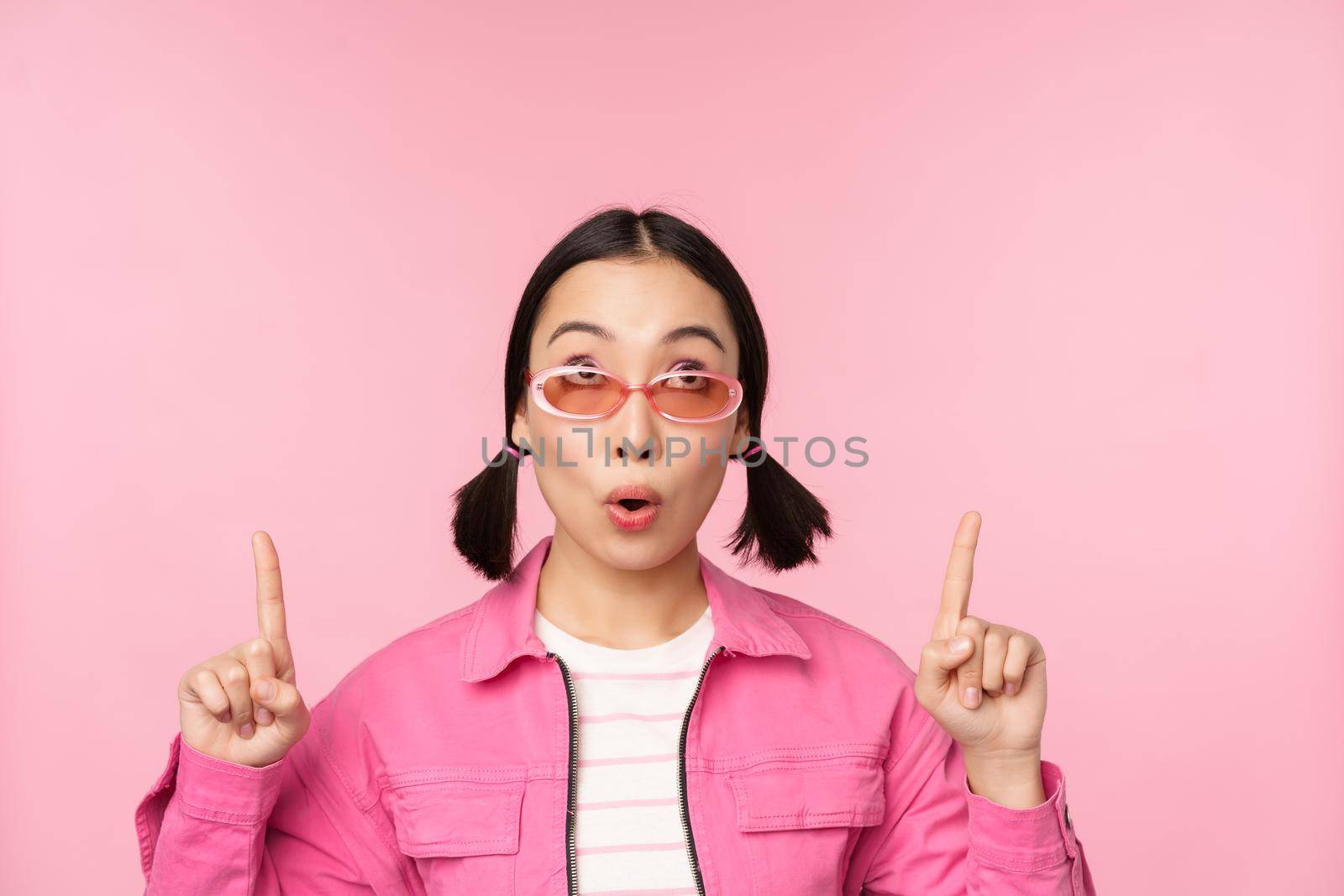 Close up portrait of asian woman gasping, looking surprised, pointing finger at banner, advertisement, standing over pink background.