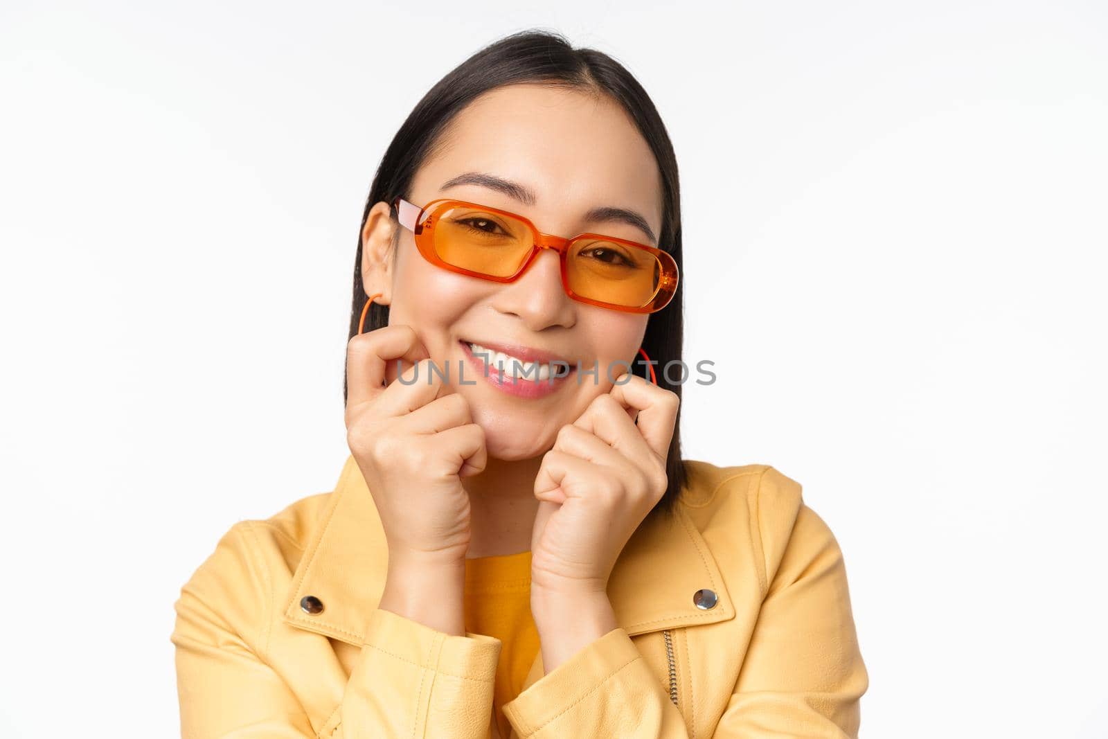 Close up portrait of trendy asian woman in sunglasses, touching her face, looking romantic, smiling at camera, standing over white background.