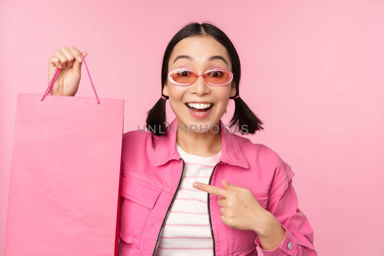 Shopping. Stylish asian girl in sunglasses, showing bag from shop and smiling, recommending sale promo in store, standing over pink background by Benzoix