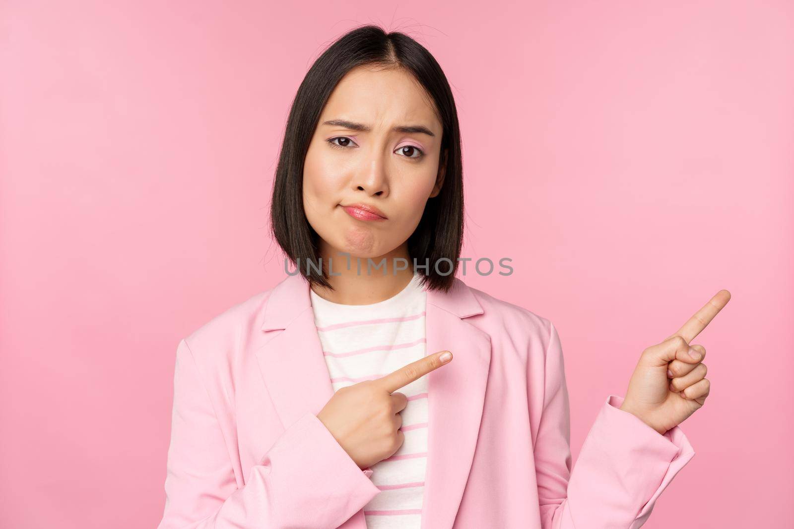 Skeptical asian businesswoman, saleswoman sulking and looking with disapproval while pointing fingers right, showing bad info, upsetting news, standing over pink background by Benzoix