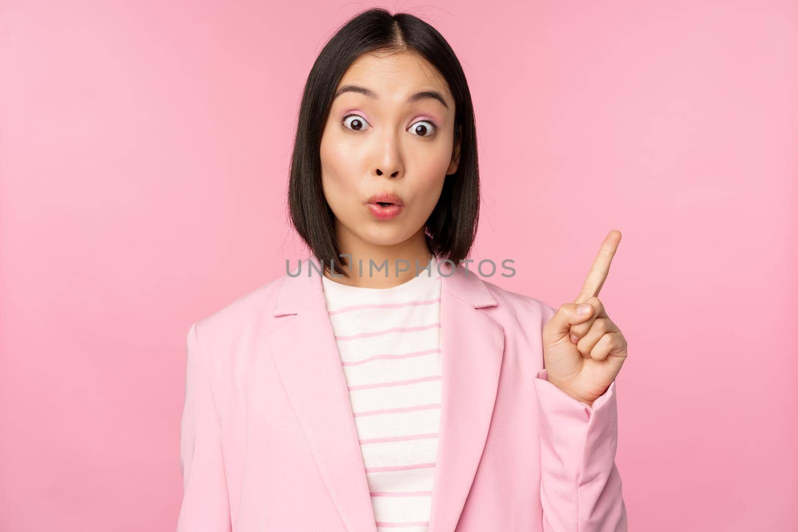 Got an idea. Young asian saleswoman, office manager raising finger, suggesting, wearing suit, posing against pink studio background by Benzoix
