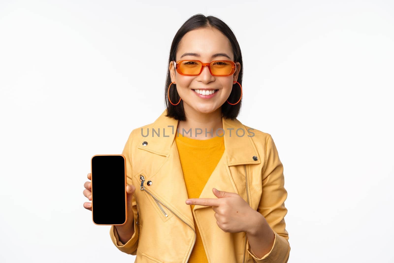 Portrait of stylish korean girl in sunglasses, smiling, pointing finger at smartphone screen, showing mobile phone application, standing over white background by Benzoix