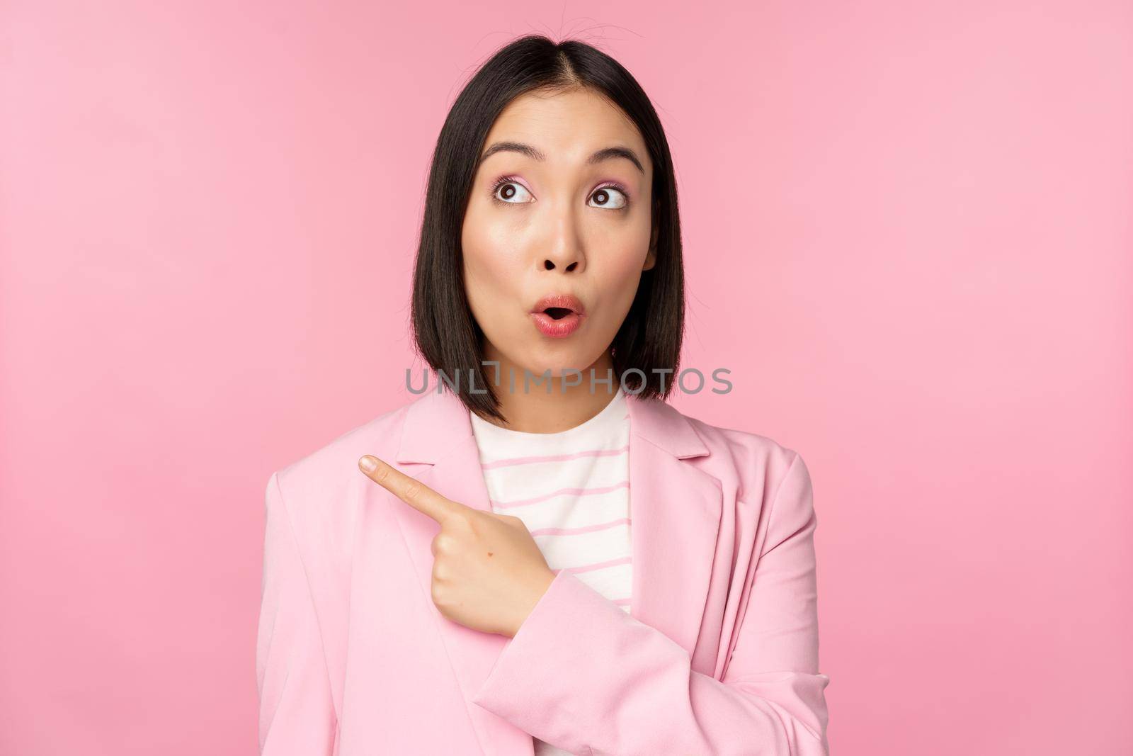 Portrait of saleswoman, korean businesswoman pointing and looking left with surprised, intrigued face expression, posing in suit over pink background.