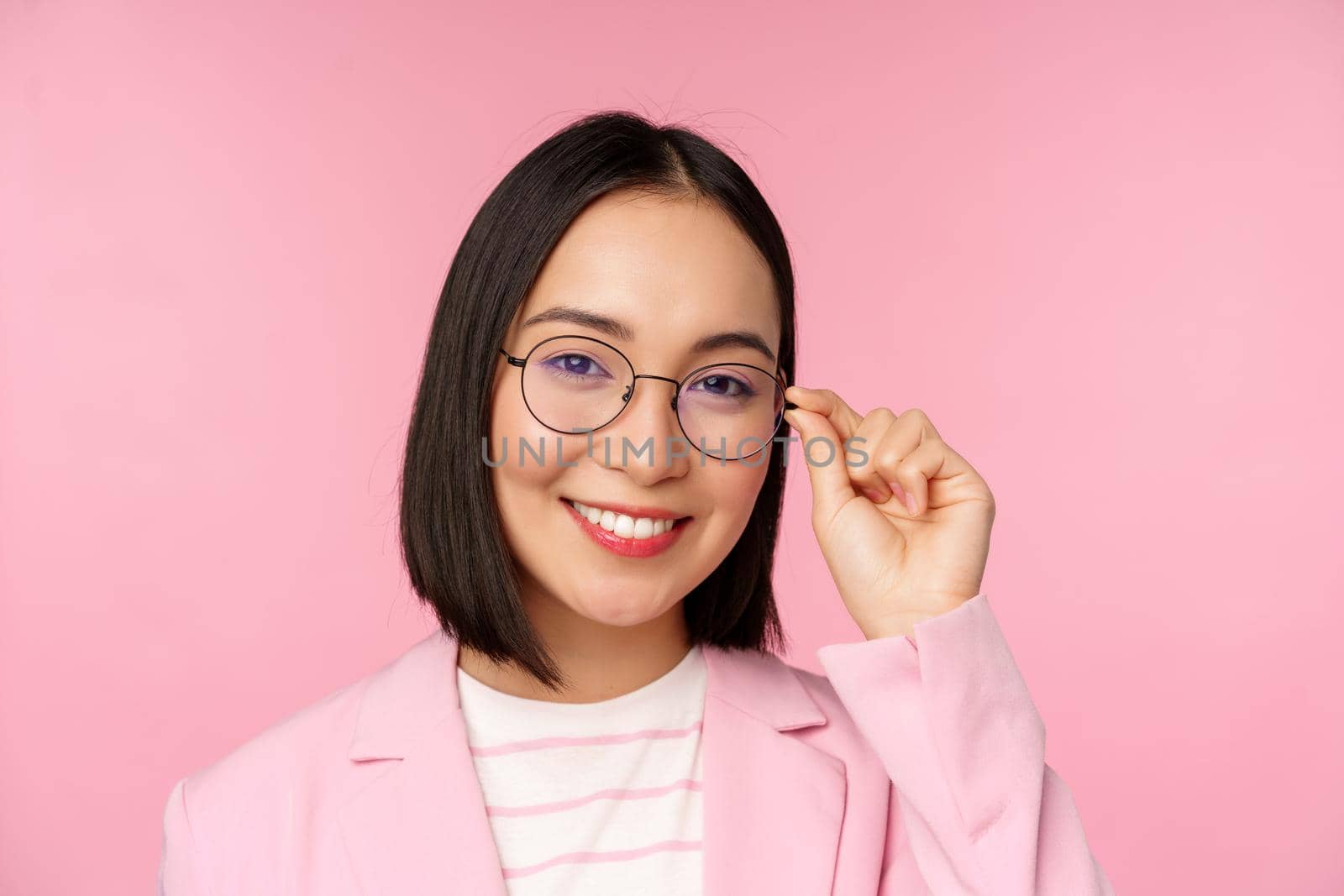 Successful asian businesswoman in glasses, smiling and looking professional at camera, wearing suit, standing over pink background by Benzoix