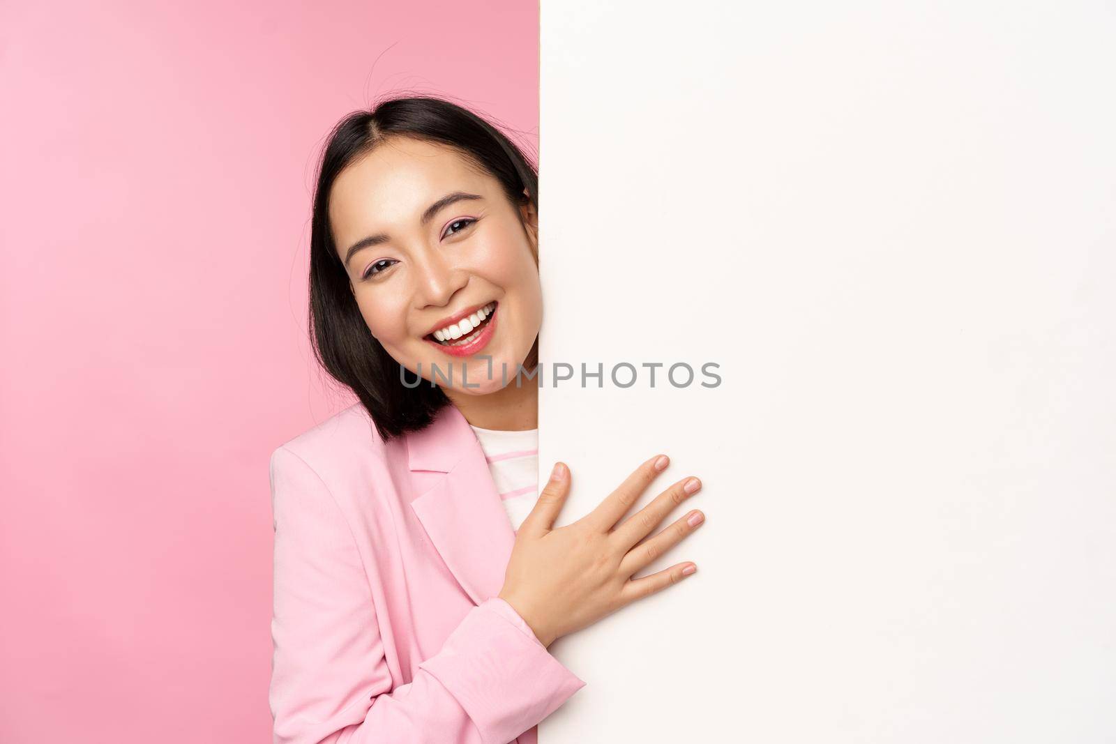 Portrait of young japanese business woman, corporate lady in suit pointing on wall with chart, showing diagram or advertisement on empty copy space, pink background by Benzoix