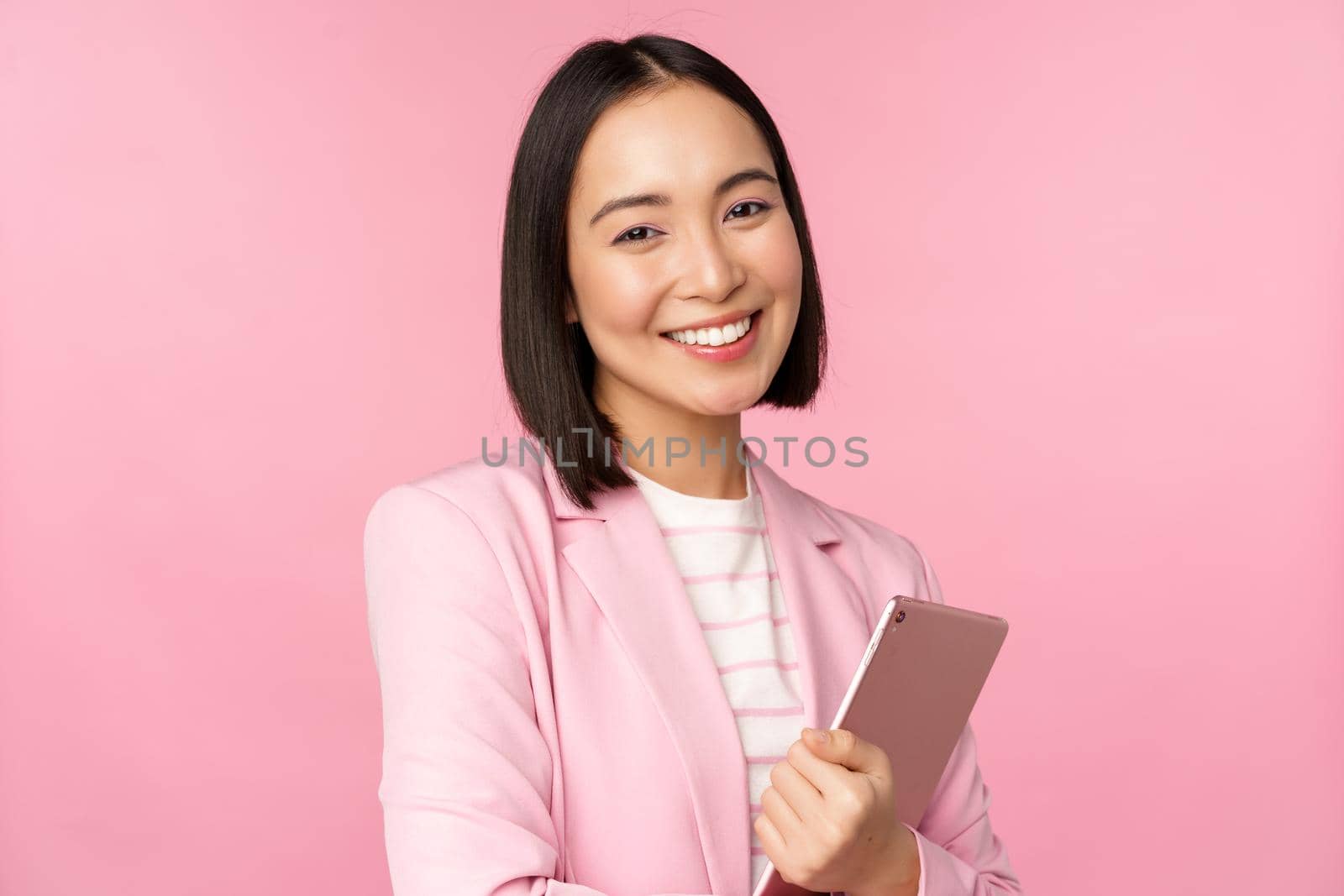 Professional smiling asian businesswoman, standing with digital tablet, wearing suit for office work, looking confident and happy, posing against pink background by Benzoix