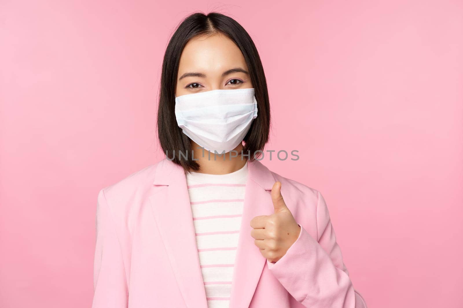 Asian businesswoman in suit and medical face mask, showing thumbs up, recommending wearing personal protective equipment in office during covid-19 pandemic, pink background.