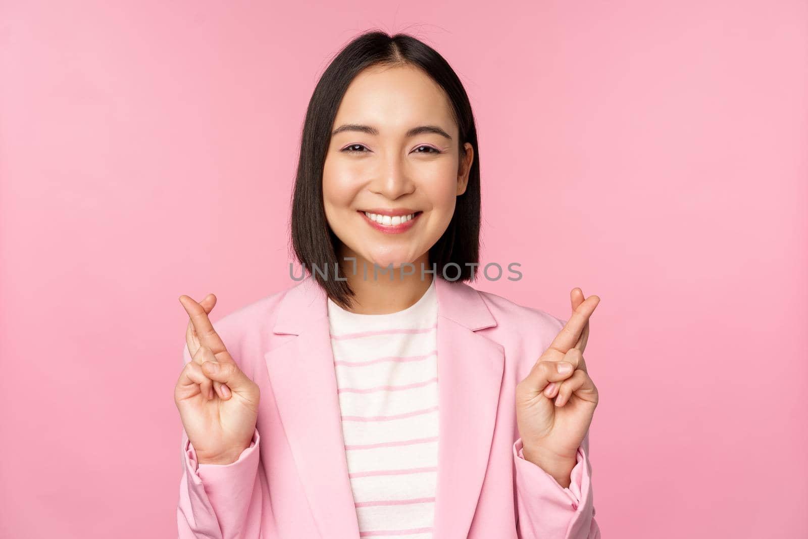 Happy asian businesswoman cross fingers for good luck, wishing, praying and hoping, smiling at camera, standing in suit over pink background by Benzoix