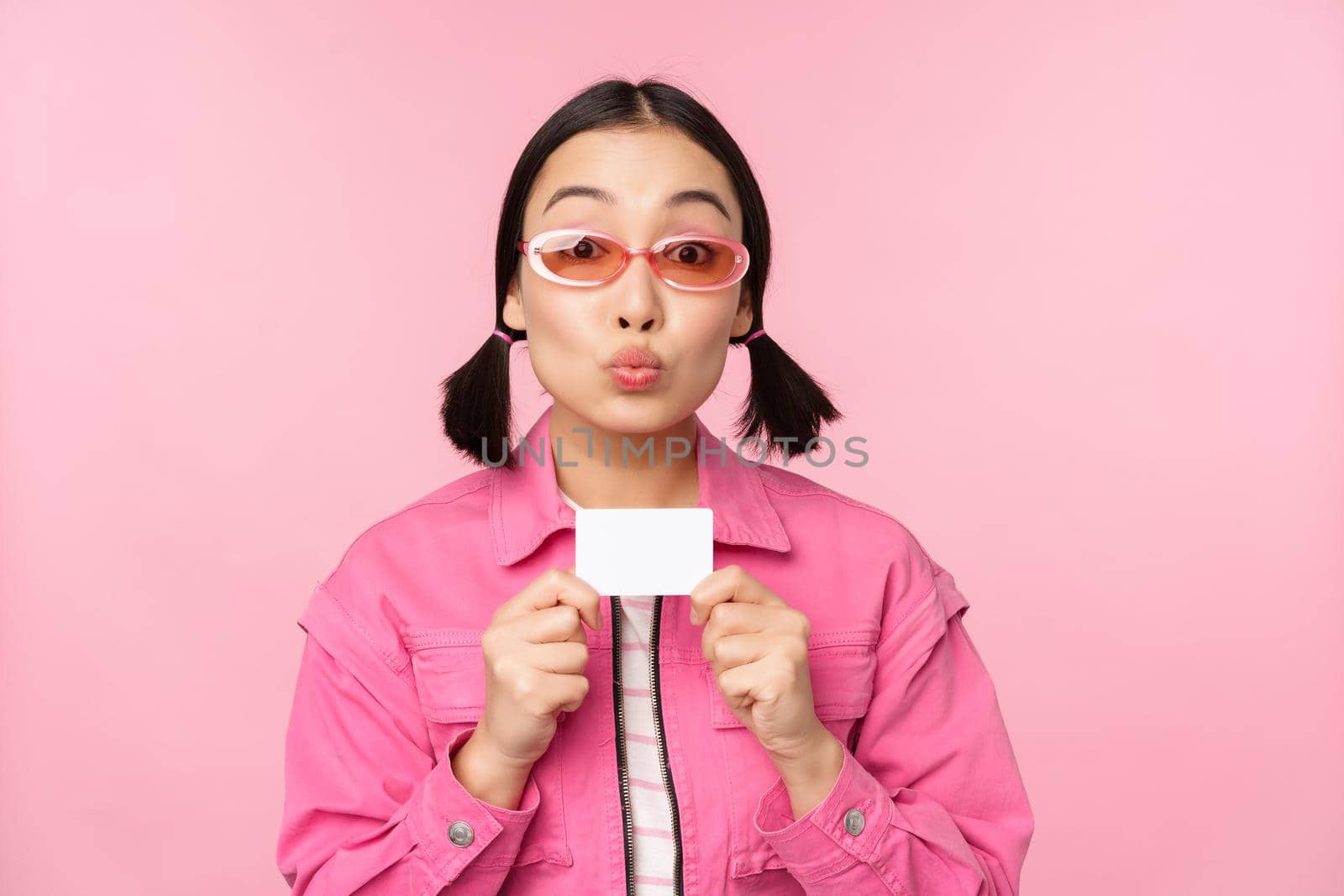 Portrait of stylish, modern asian girl, shows discount, credit card and look pleased, paying contactless, concept of shopping, standing over pink background by Benzoix