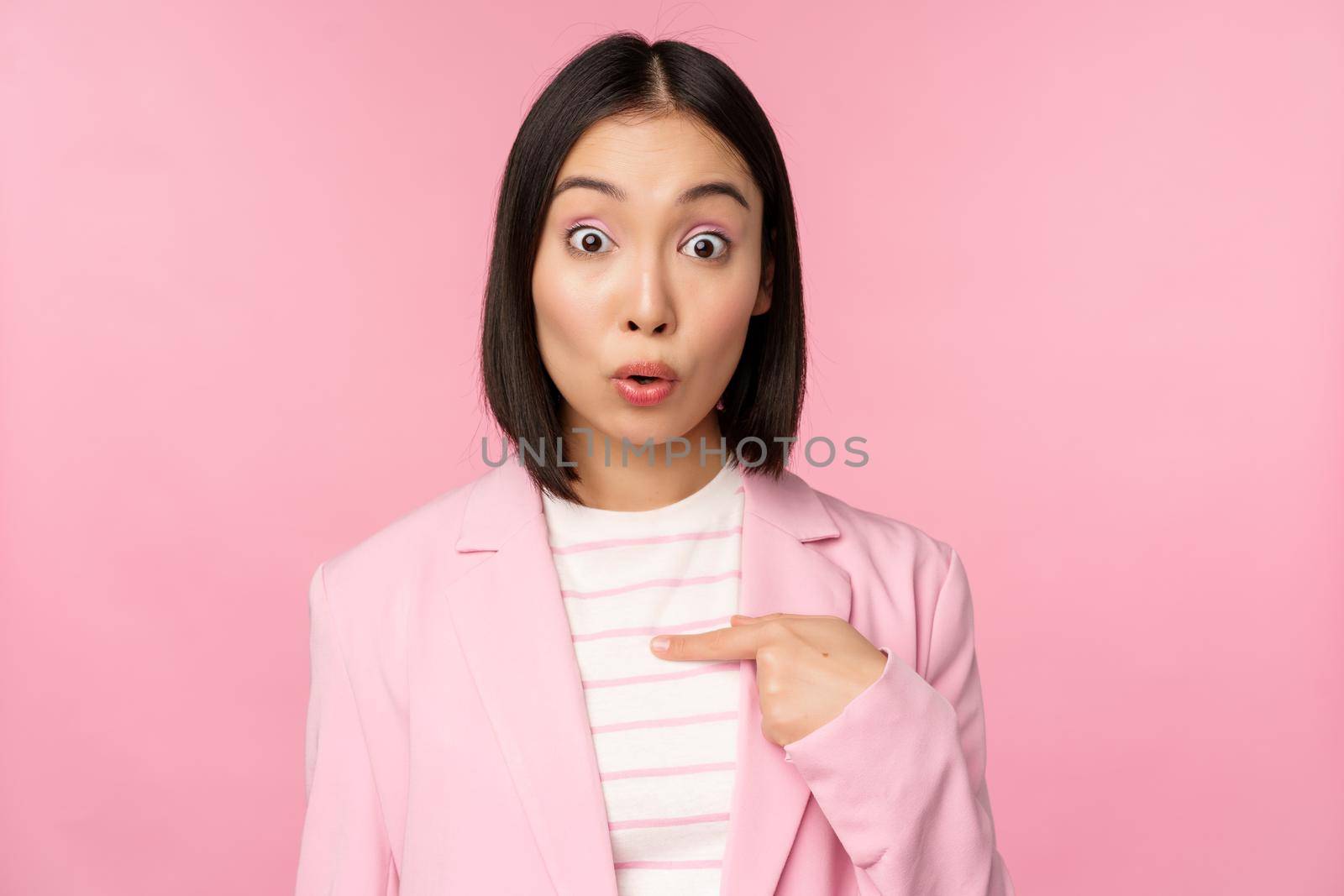 Portrait of asian businesswoman reacts surprised, points at herself with disbelief on face, posing in suit against pink background by Benzoix