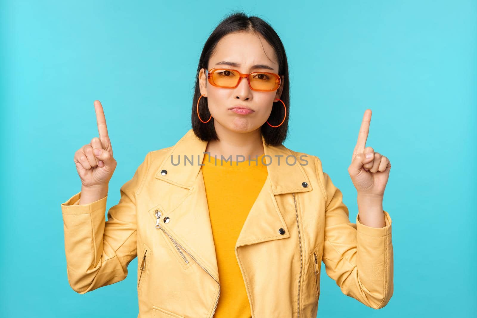 Upset, gloomy korean woman in sunglasses, points fingers up, looks disappointed while shows banner on top, stands over blue background.