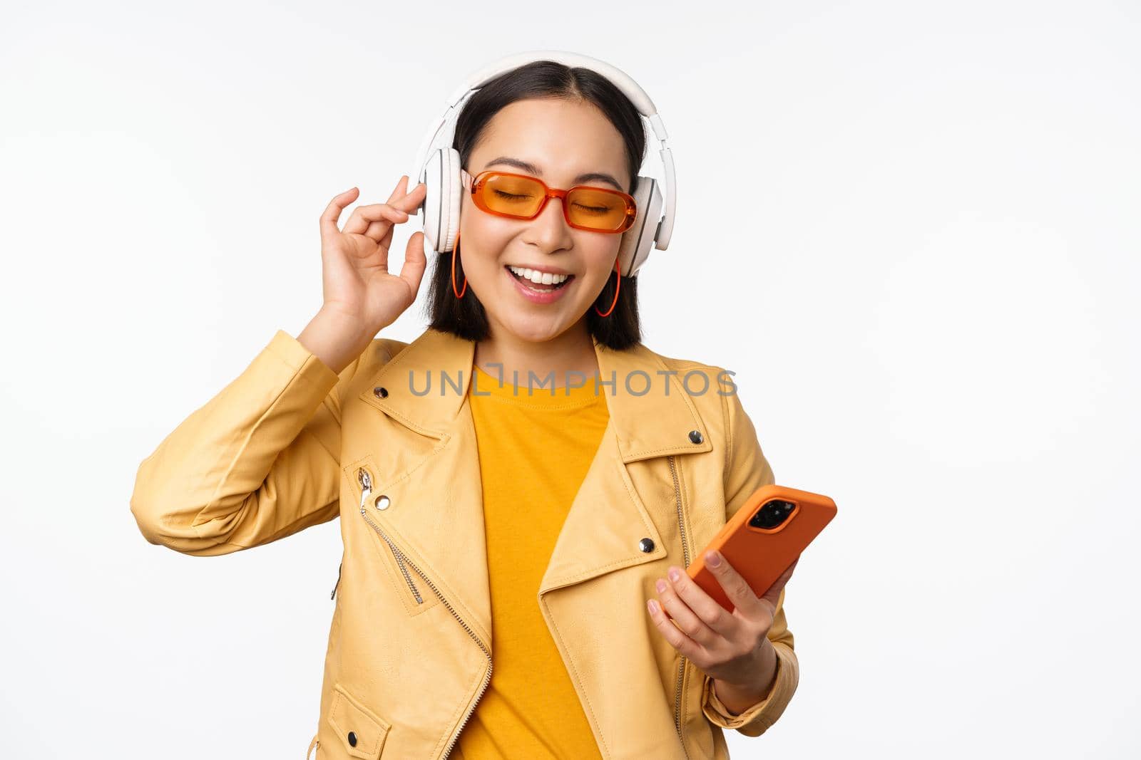 Happy stylish asian woman in sunglasses, listening music in headphones, holding smartphone, singing and dancing, standing over white background by Benzoix
