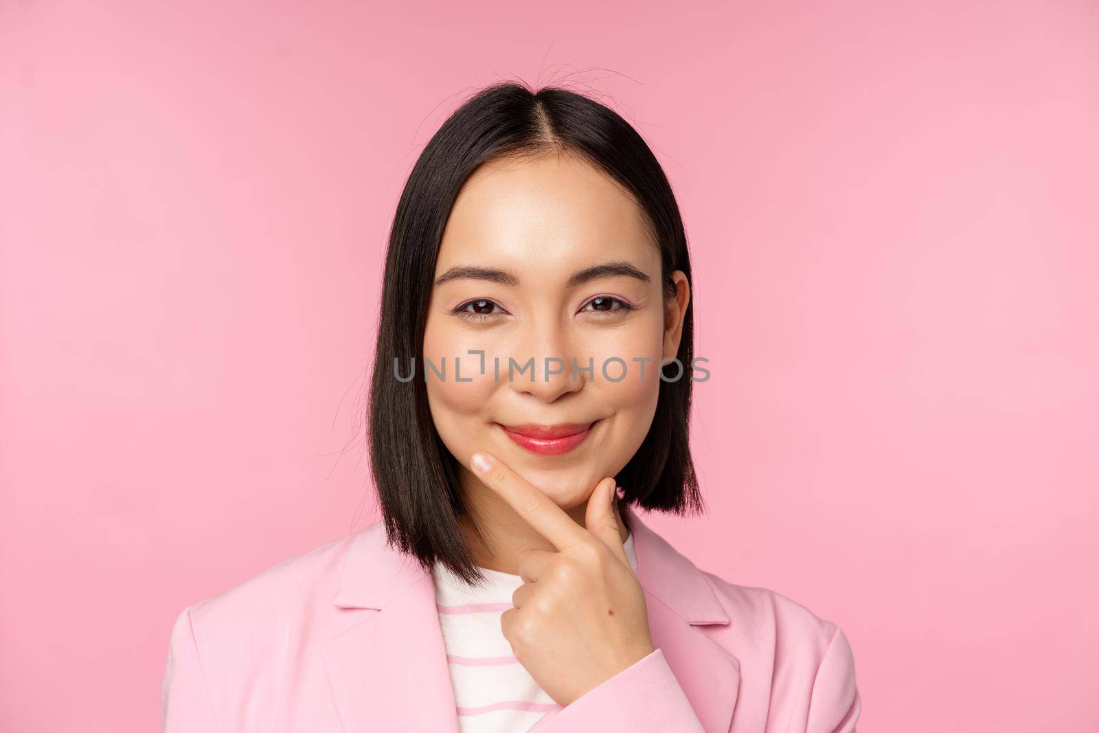 Close up portrait of smiling asian working lady in suit, businesswoman looking thoughtful, thinking or deciding smth, standing over pink background.