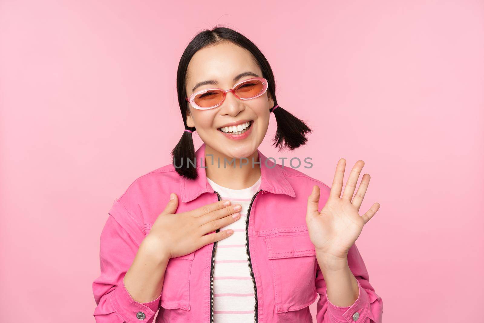 Portrait of friendly asian teen girl in sunglasses saying hello, waves her hand and smiles, greets you, hi gesture, stands over pink background by Benzoix