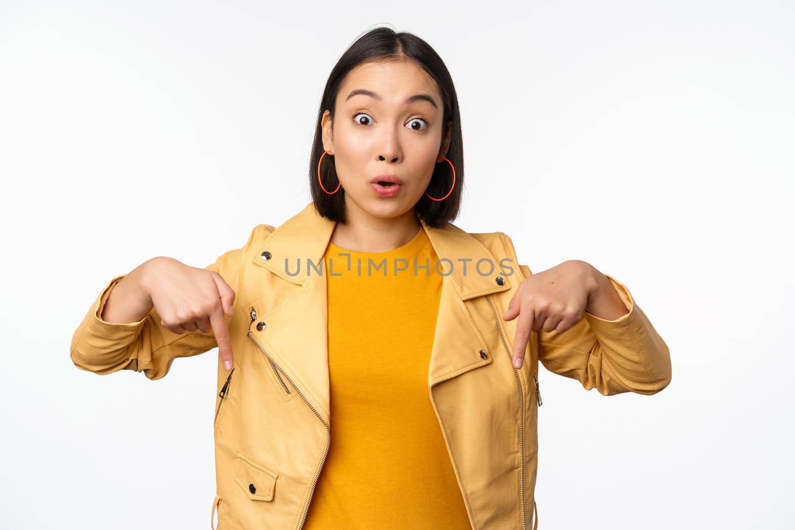 Portrait of surprised asian girl, looking amazed and impressed, pointing fingers down, showing banner with shocked face expression, white background by Benzoix