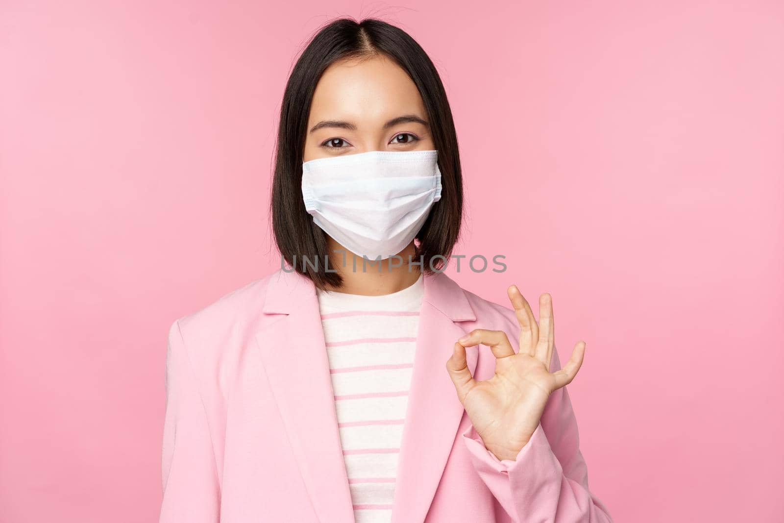 Portrait of asian businesswoman in medical face mask, showing okay sign, wearing suit, work rules during covid-19 pandemic, standing over pink background.