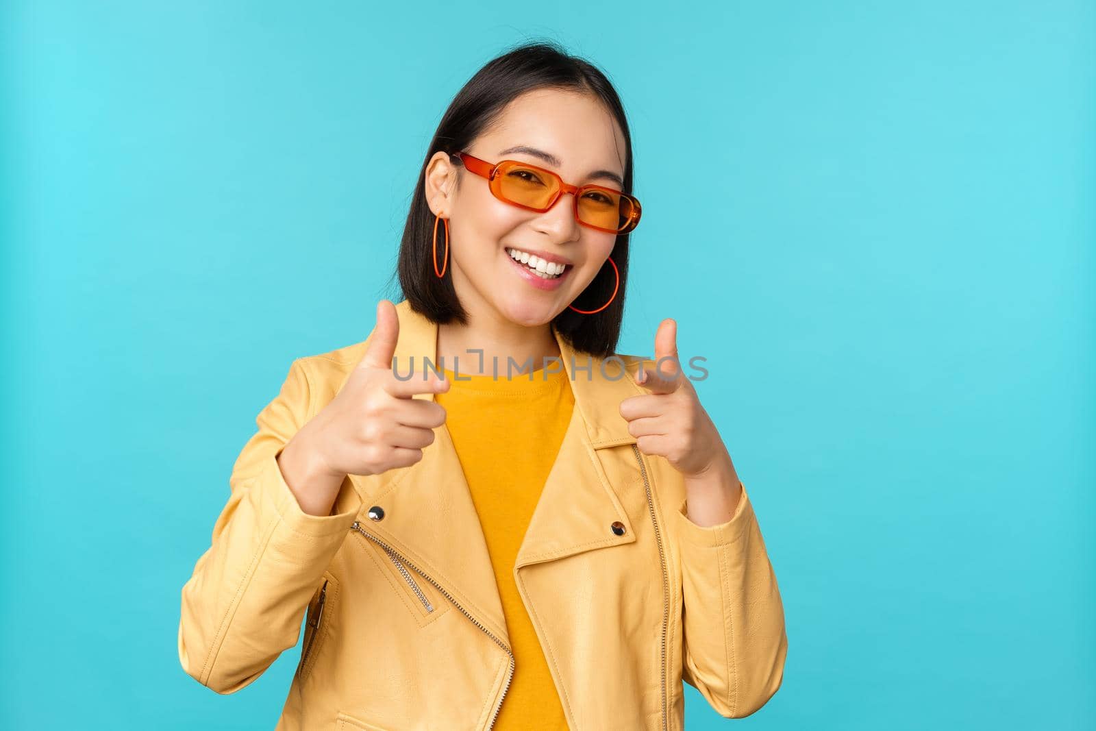 Happy young stylish chinese girl in sunglasses, points fingers at camera with pleased smile, choosing you, congratulating, standing over blue background.