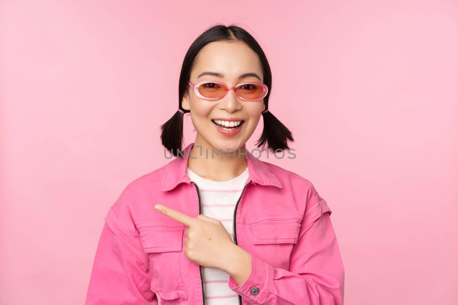 Portrait of smiling asian girl in stylish outfit, sunglasses, pointing finger left, showing advertisement, banner, standing over pink background. Copy space