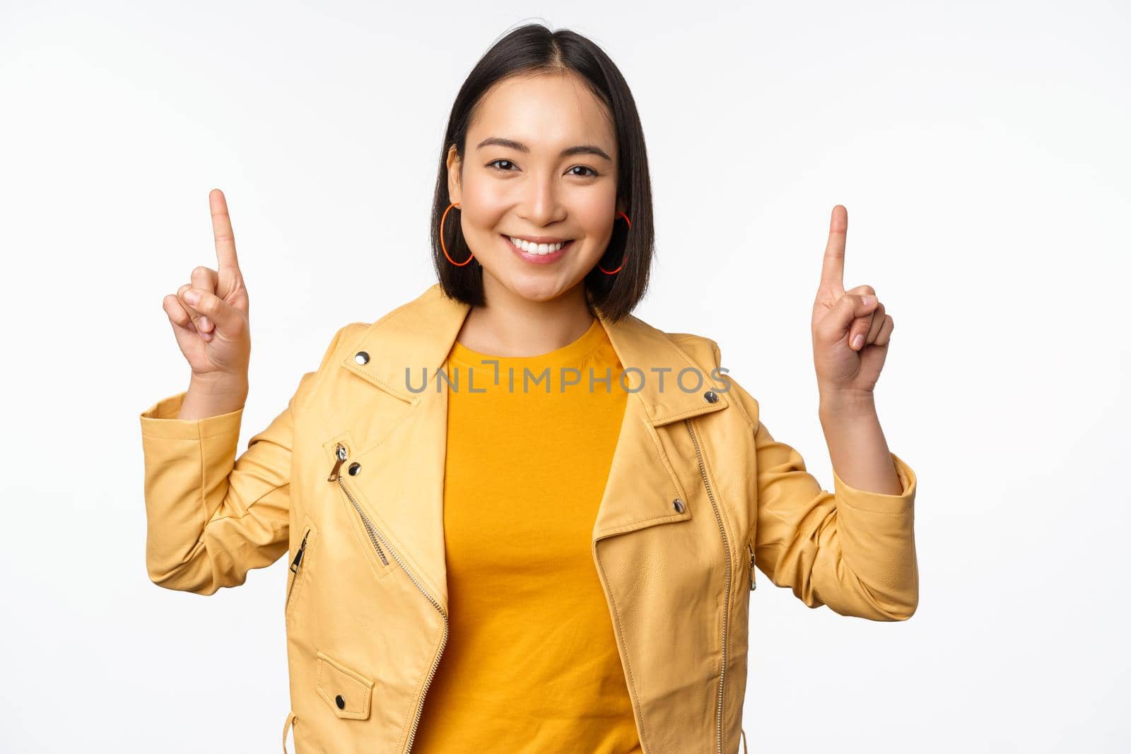 Enthusiastic asian girl pointing fingers up, showing advertisement on top, smiling happy, demonstrating promo offer or banner, standing over white background.