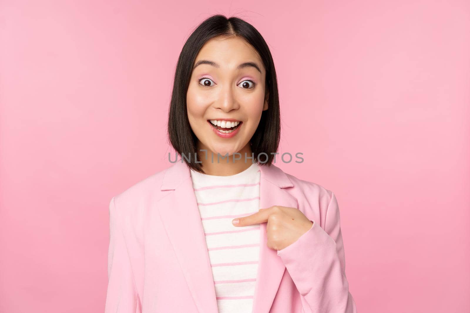 Portrait of young asian businesswoman with surprised, excited face expression, pointing finger at herself, standing in suit over pink background.
